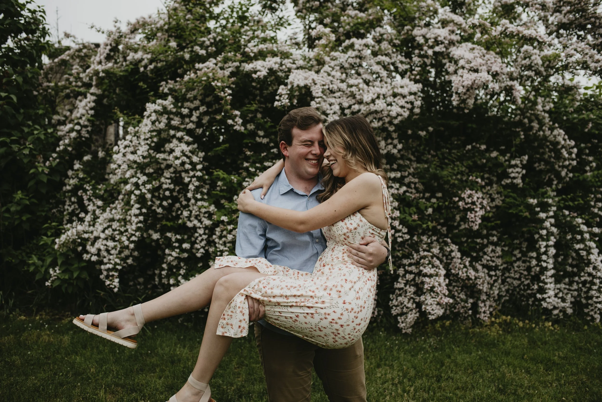 A man holding a woman in his arms in front of blooming white flowers. They are smiling and appear happy.