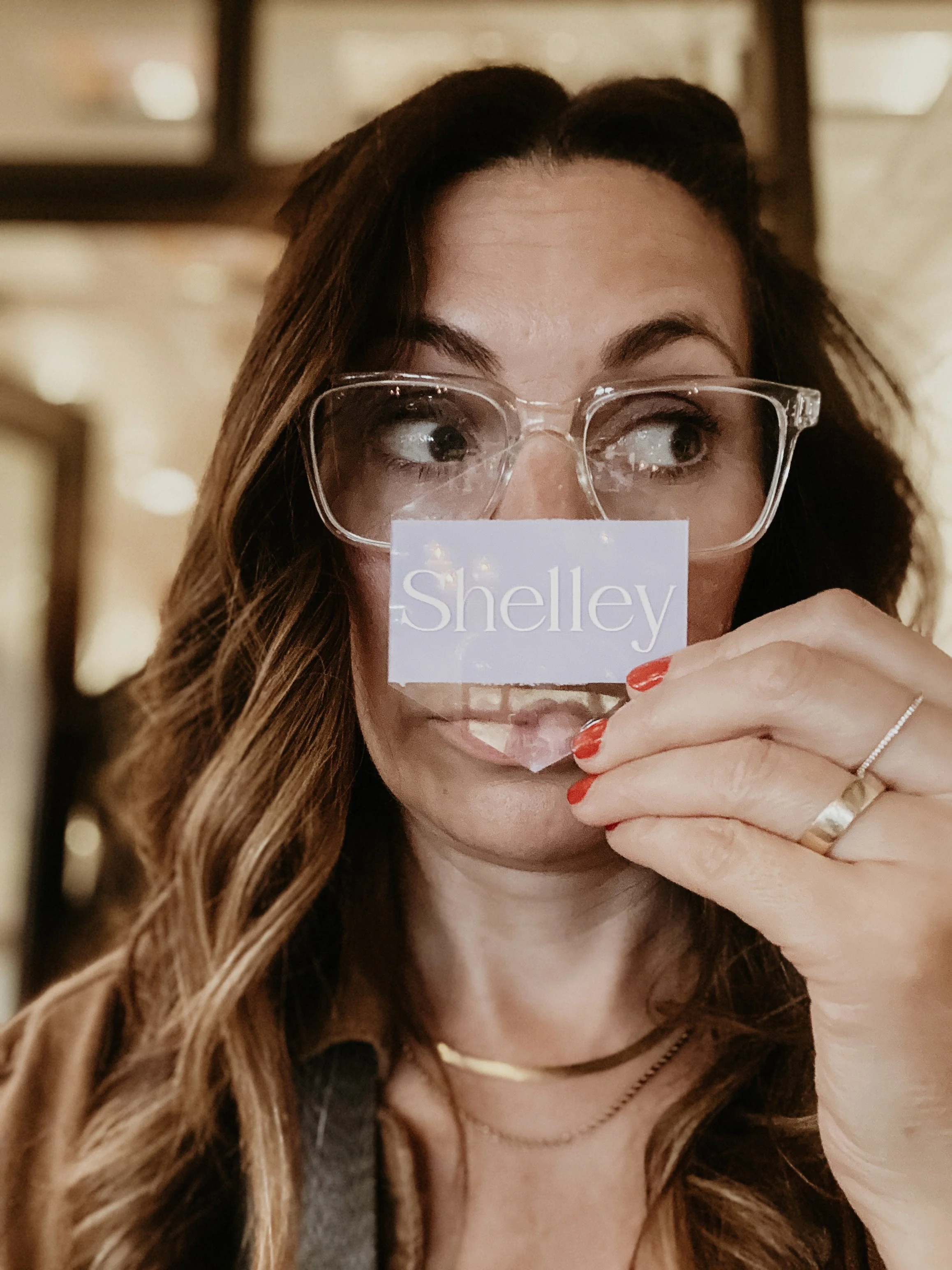 A woman with long wavy brown hair wearing glasses partially covering her face with a sign that says "Shelley." She has red nail polish, multiple rings, and a necklace. The background is indoors with warm lighting.