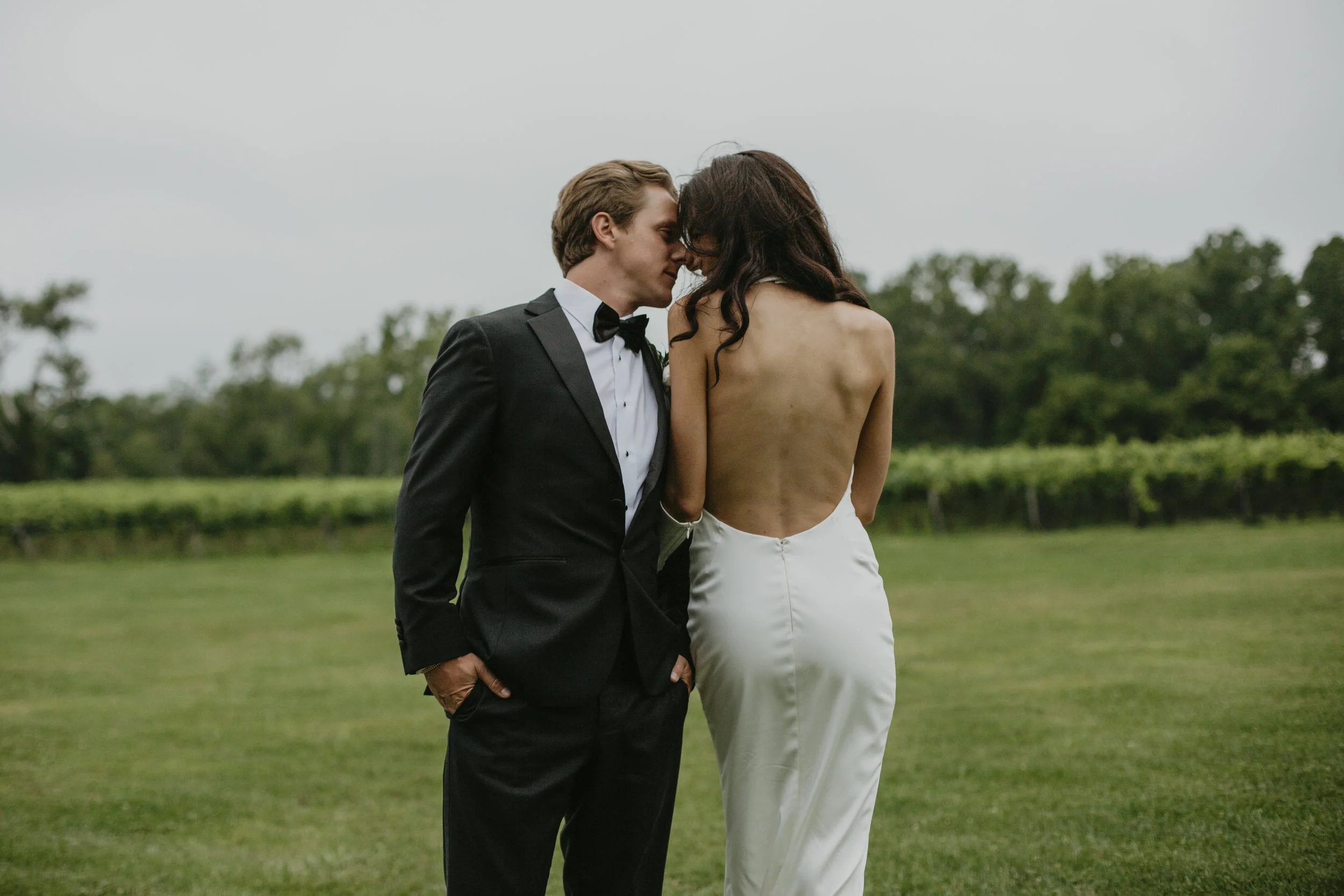 A couple in wedding attire, with the groom in a tuxedo and the bride topless in white pants, standing close together outdoors on a grassy field with trees and overcast sky in the background.