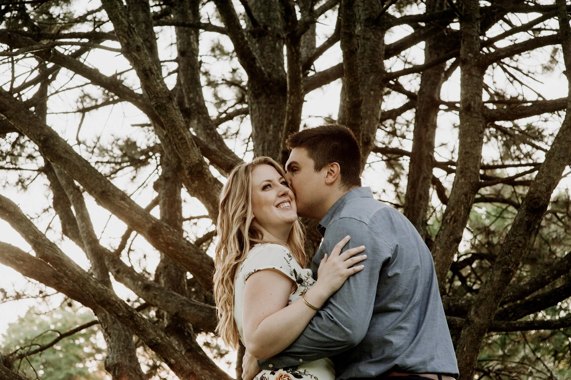A young couple embracing outdoors in front of a large tree with many branches.