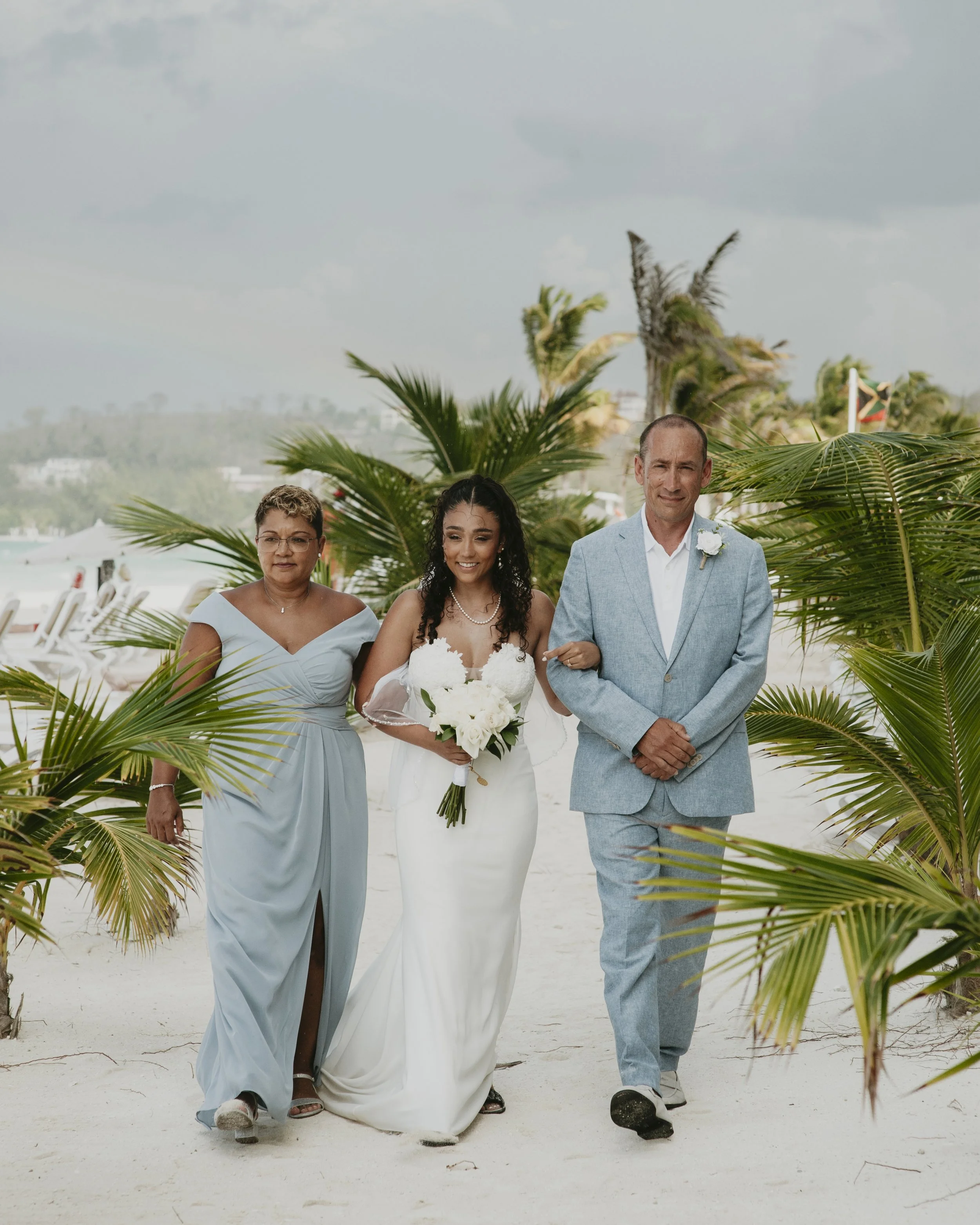 A bride is walking down a sandy beach aisle with two older adults, surrounded by palm trees, during a wedding ceremony.