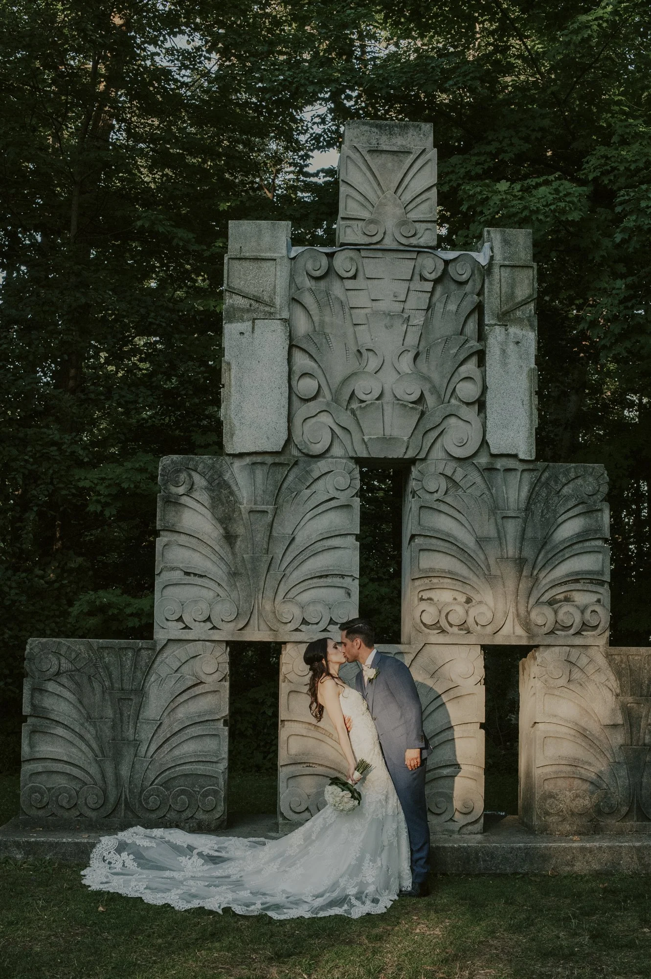 A bride and groom share a kiss in front of a large decorative stone structure with intricate carvings, surrounded by trees during a wedding photoshoot.
