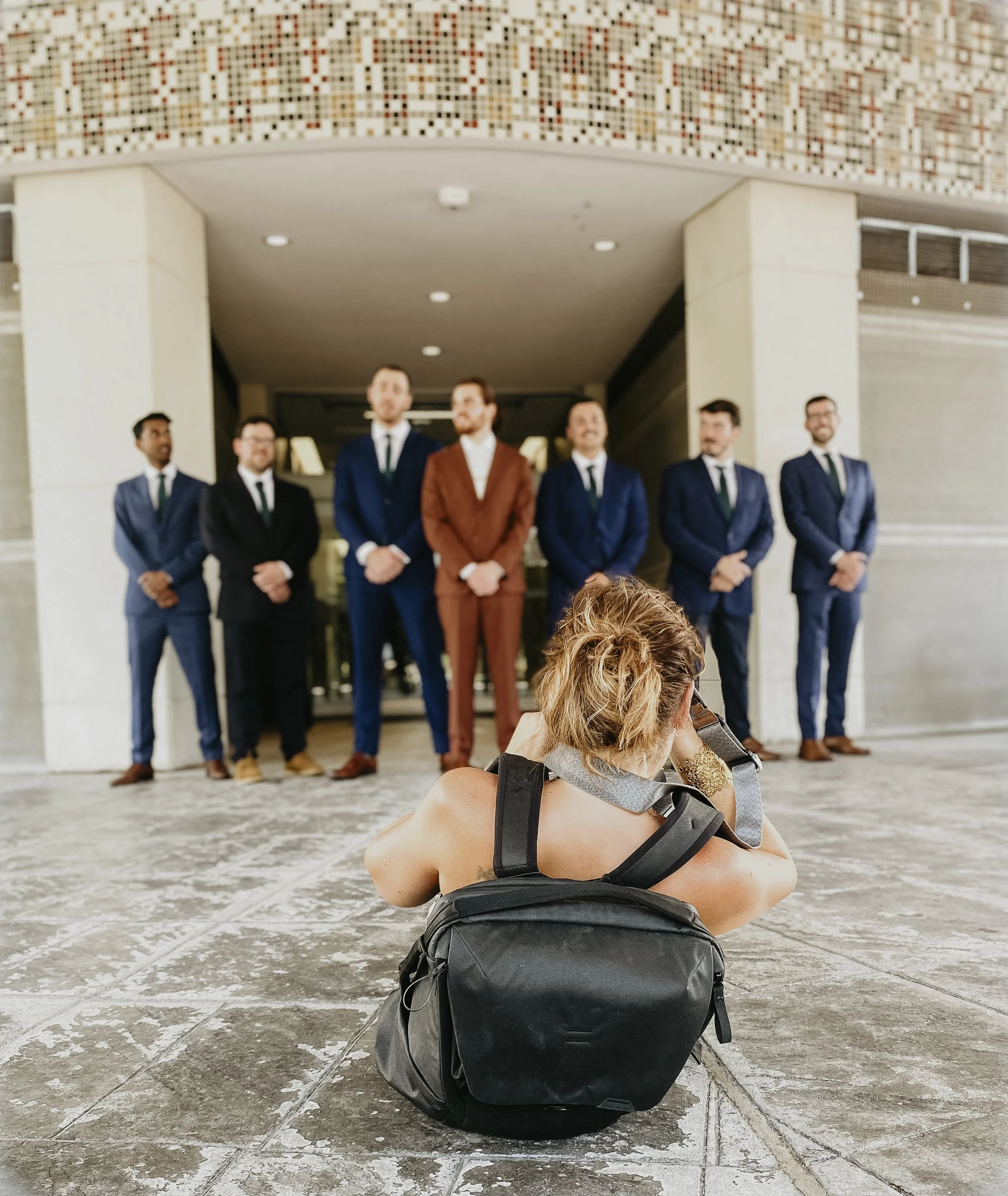 A woman with curly hair resting on a backpack, sitting on the ground outdoors near a group of men in suits standing together.