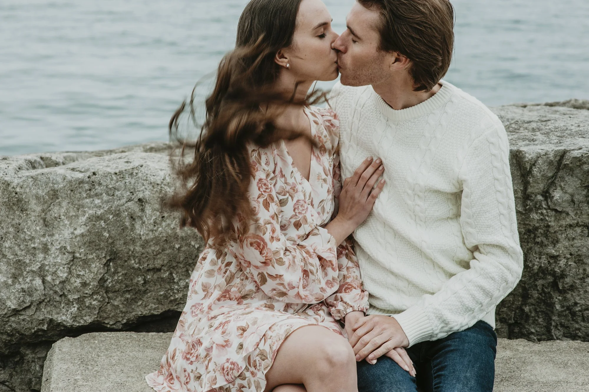 A couple sitting on a large rock near the water, sharing a kiss. The woman has long brown hair, is wearing a floral dress, and the man has brown hair, is wearing a white sweater, and dark pants.
