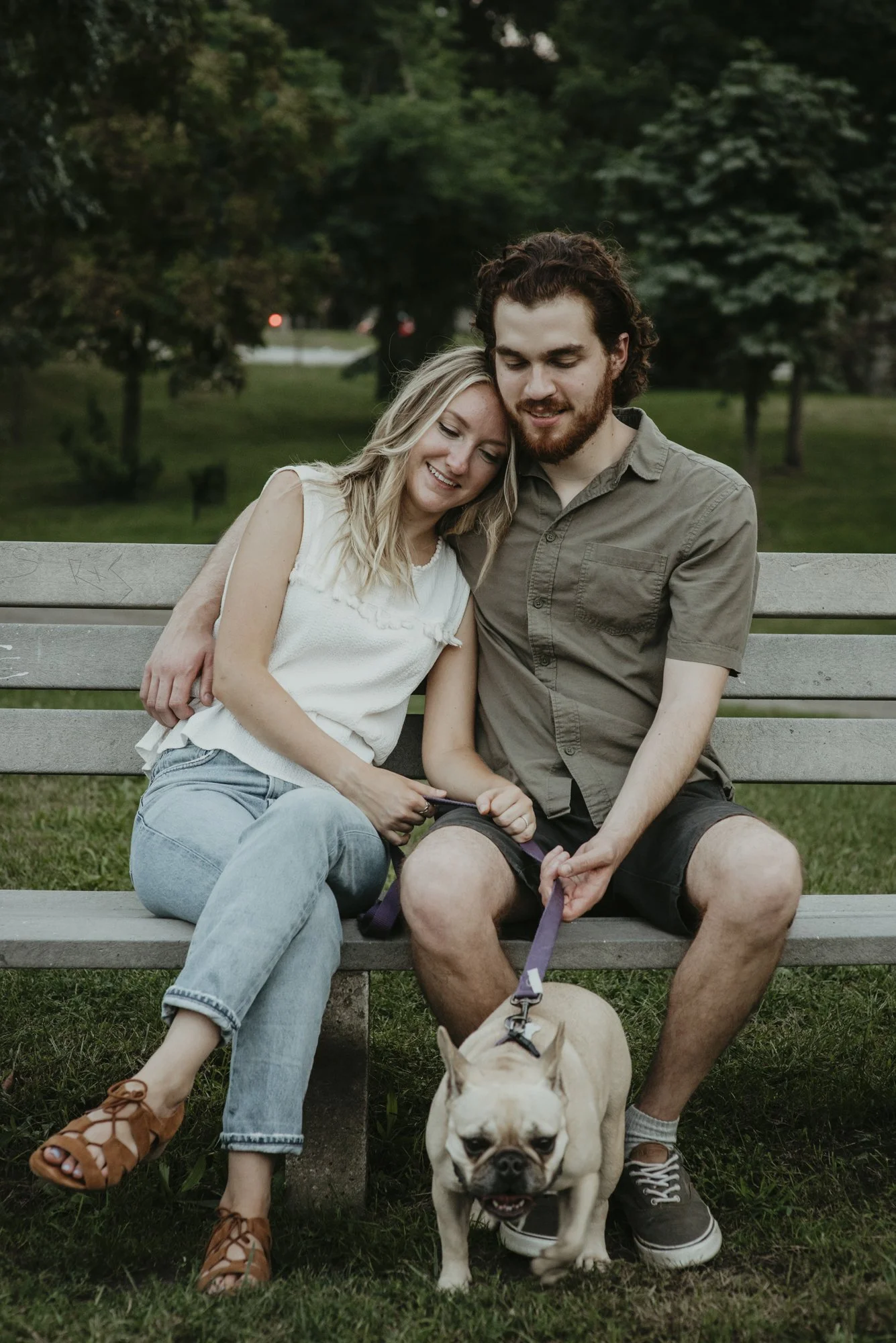 A young couple sitting on a park bench with a French Bulldog in front of them. The woman is leaning her head on the man's shoulder, both smiling, with green trees in the background at dusk.