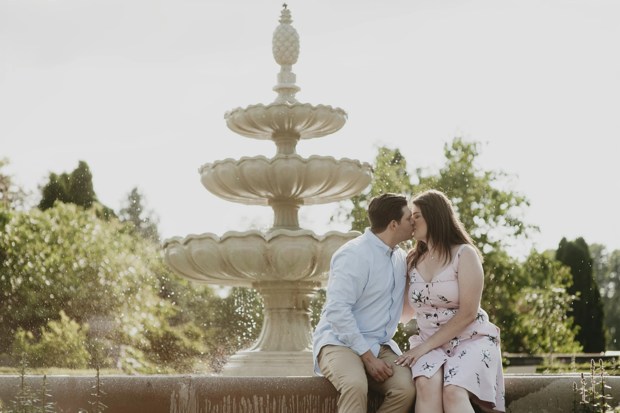 A couple kissing in front of a multi-tiered fountain outdoors on a sunny day.