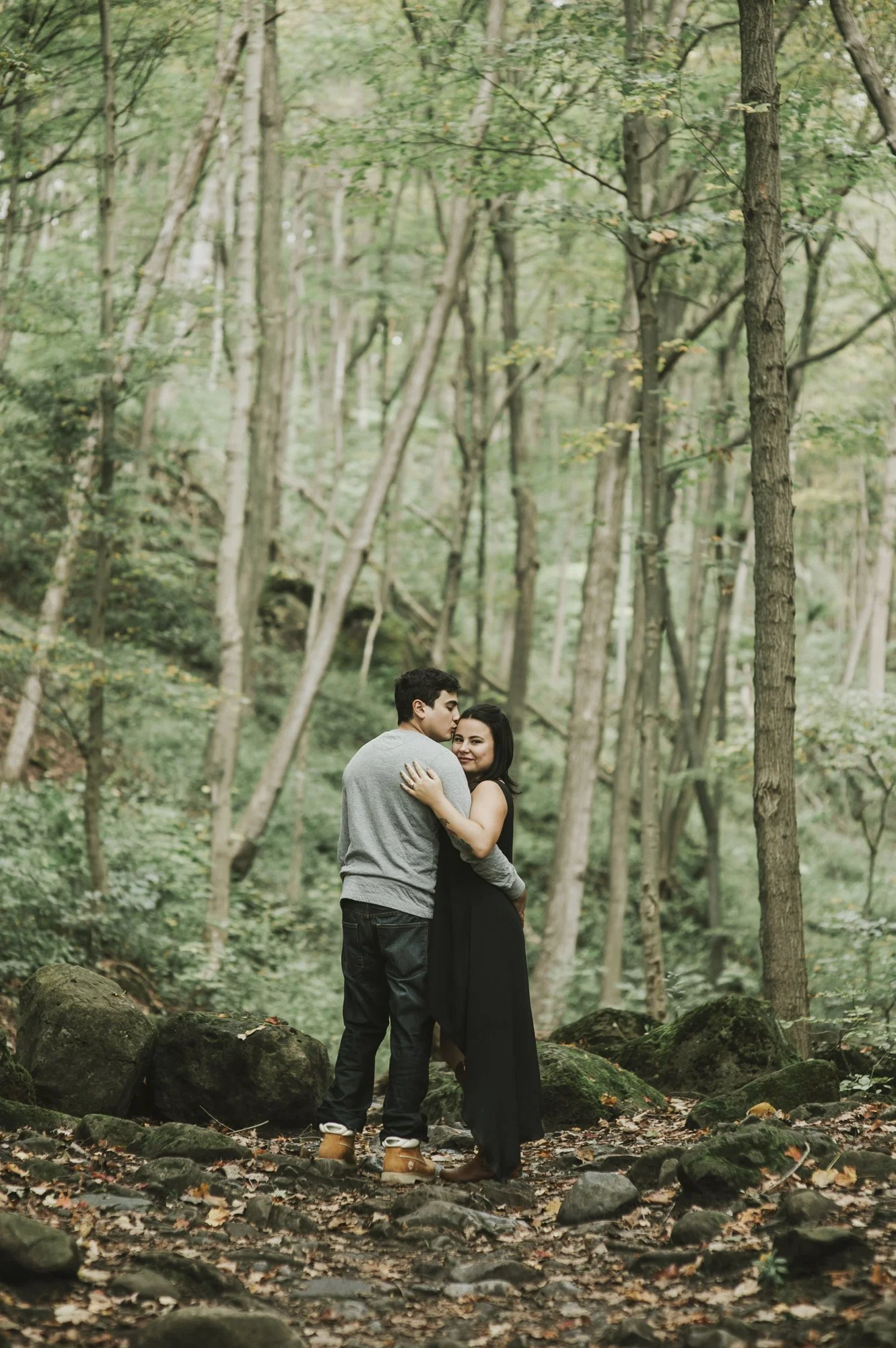 A couple embracing in a forest with trees and rocks.