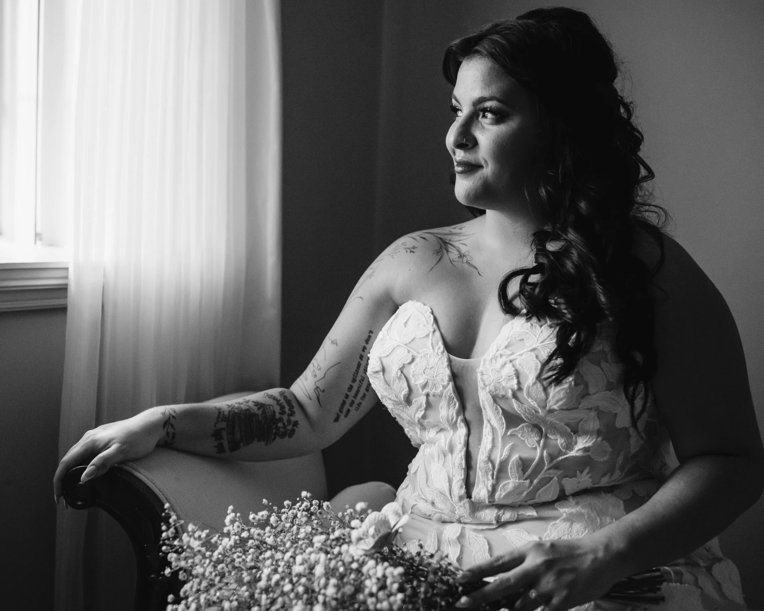 A woman with dark curly hair and tattoos on her arm sitting near a window, wearing a lace dress, holding a bouquet of flowers, looking thoughtfully outside.