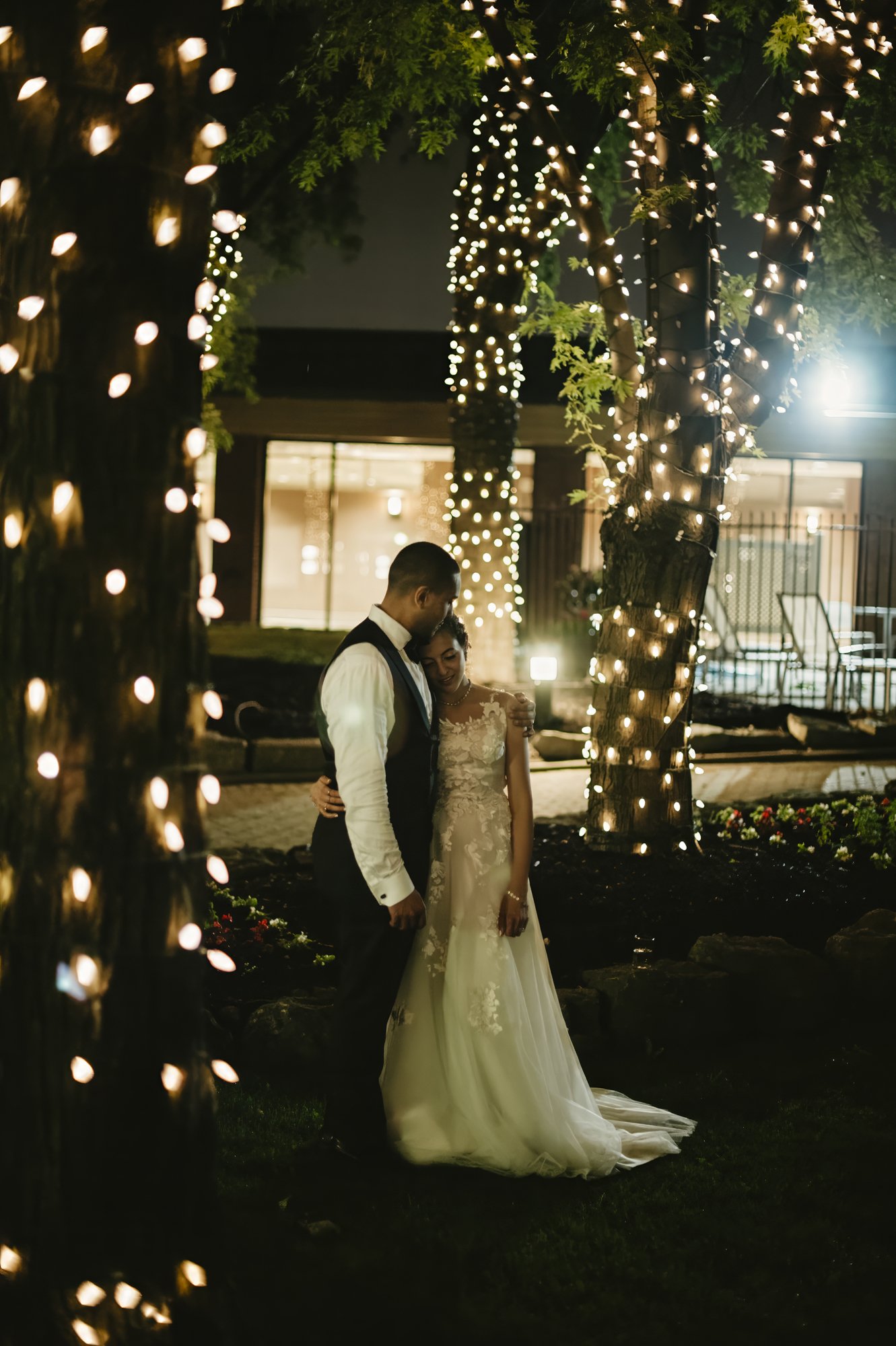 A newlywed couple stands close together in a garden at night, surrounded by trees wrapped in string lights, with a building in the background.