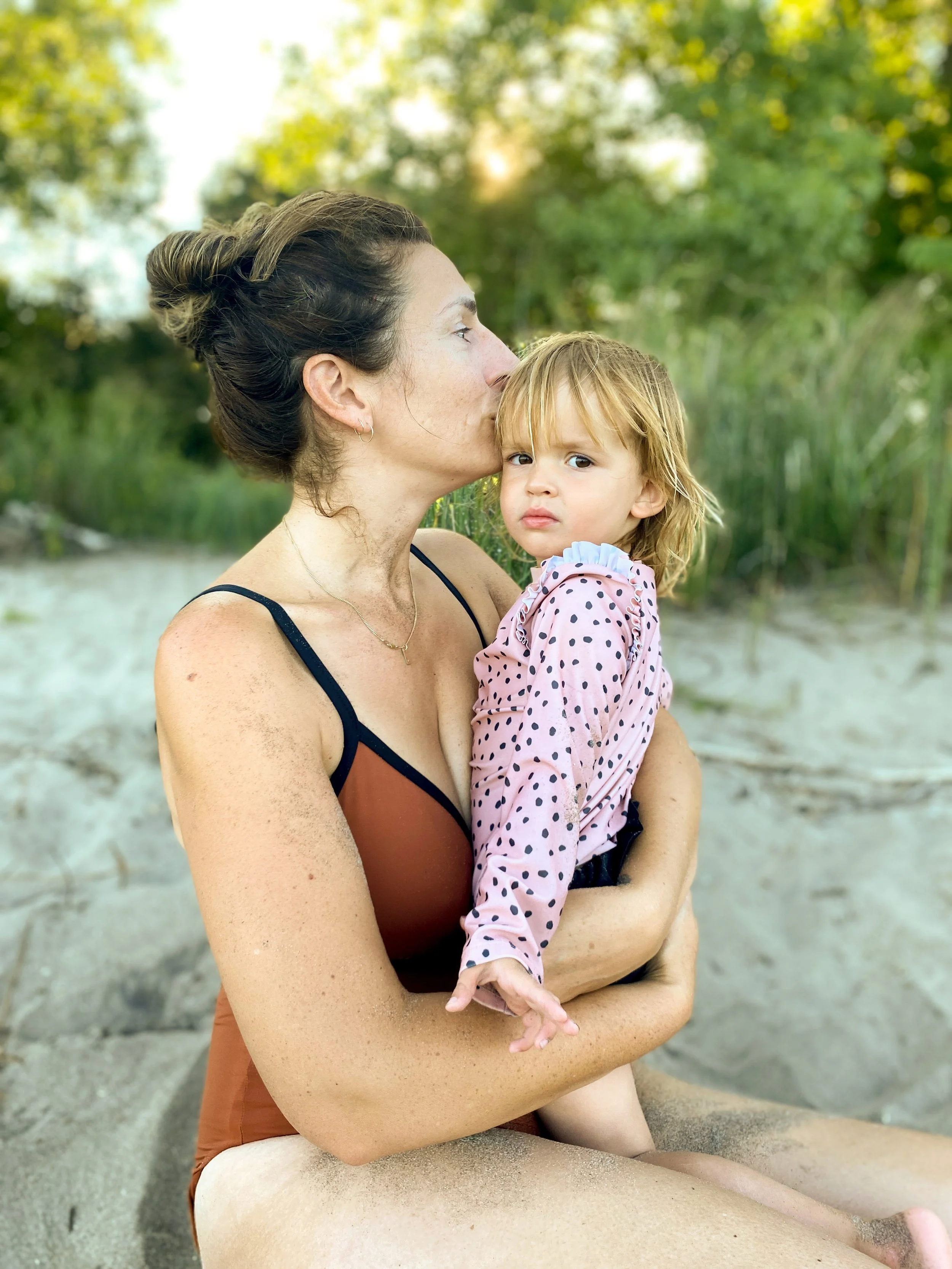A woman with dark hair in a bun holding a young girl with blonde hair on her lap, near a sandy beach with green trees in the background. The woman is kissing the girl on the forehead, and the girl is looking at the camera with a neutral expression.