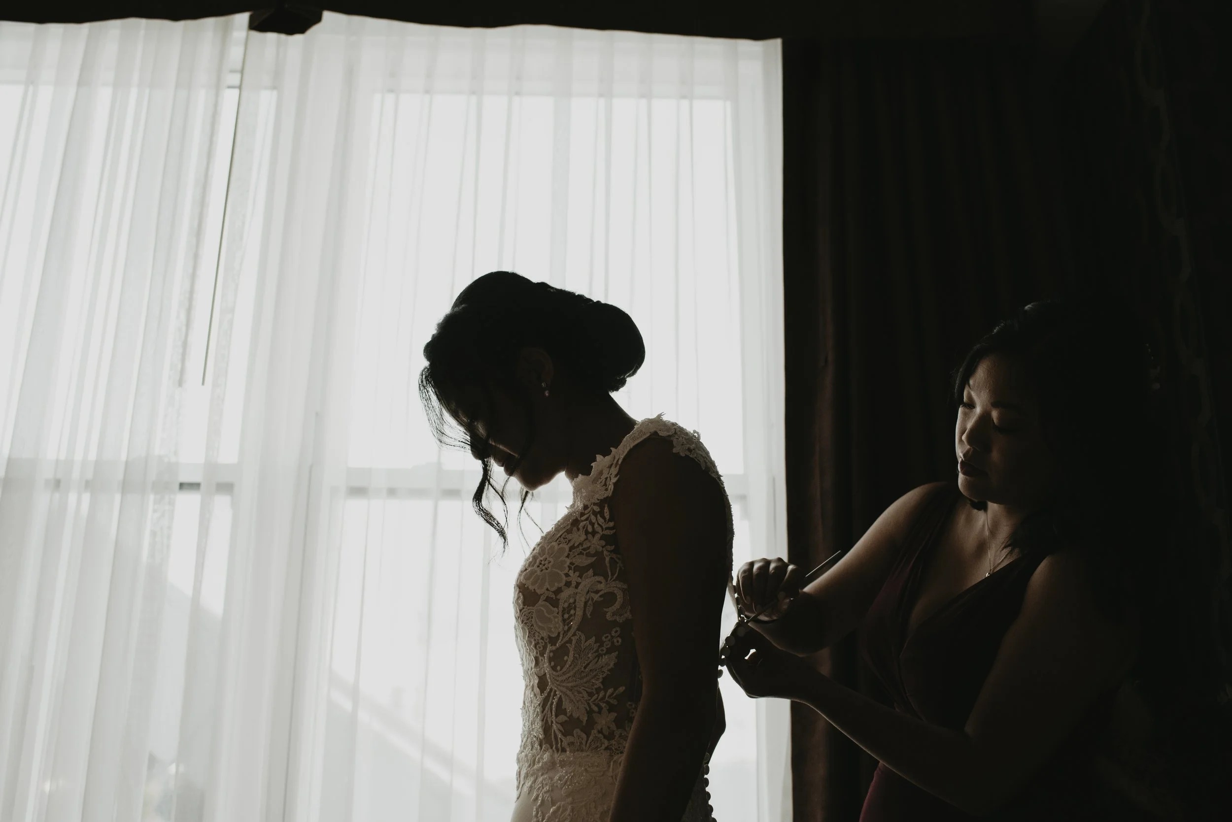 A woman in a lace wedding dress is being assisted by another woman in a dark dress as she prepares for her wedding, near a large window with sheer curtains.