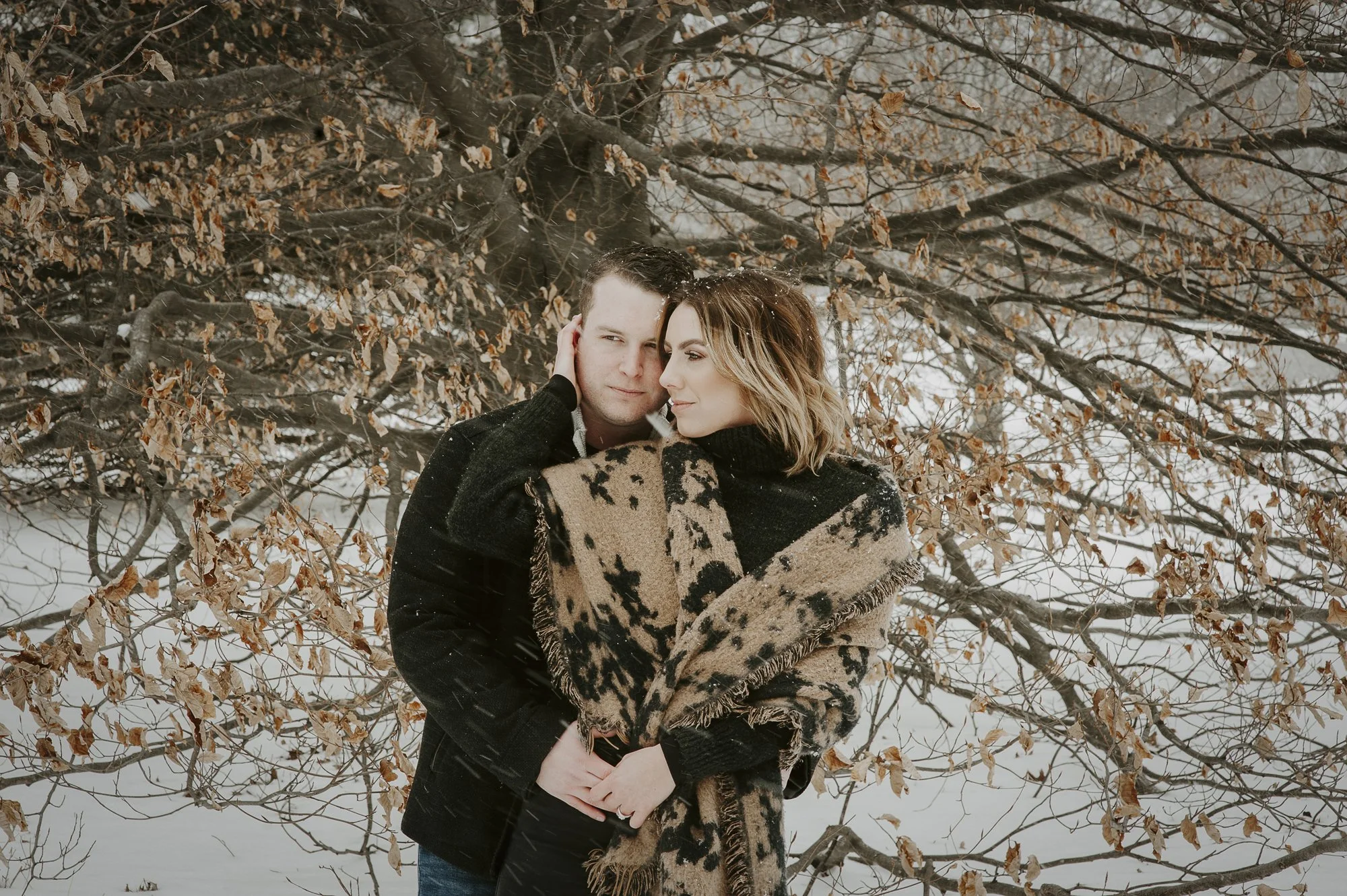 A couple standing close together outdoors in winter, with a tree with brown leaves and snow in the background. The woman is wrapped in a animal print coat and the man is wearing a dark jacket. They look at each other affectionately.