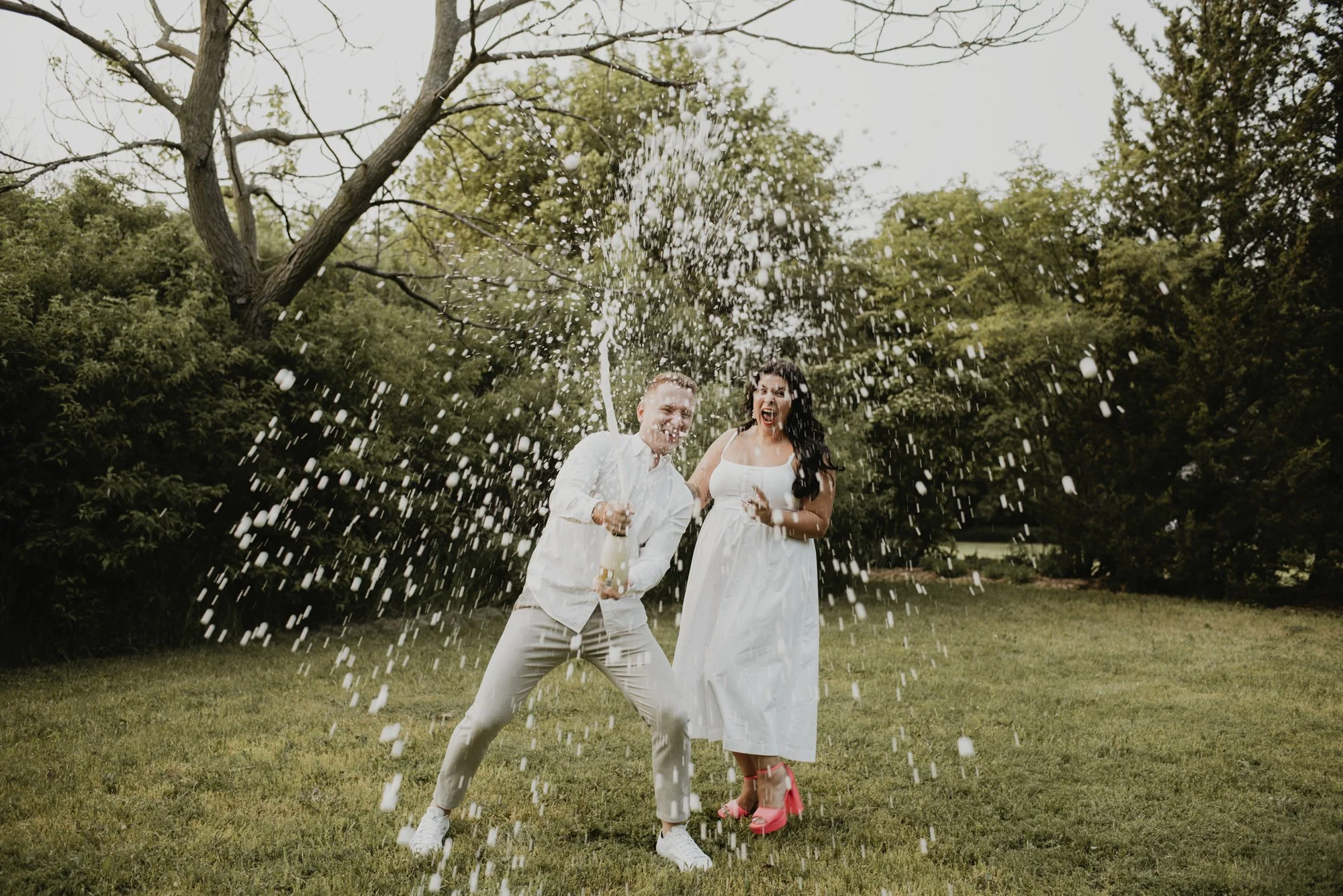 Two people, a man and a woman, in wedding attire celebrating outdoors with a bottle of champagne, splashing champagne in the air.