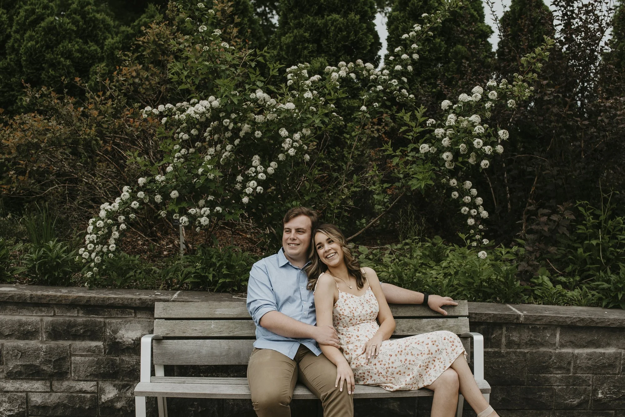 A young couple sitting on a park bench surrounded by lush greenery and white flowering bushes, smiling and enjoying each other's company.