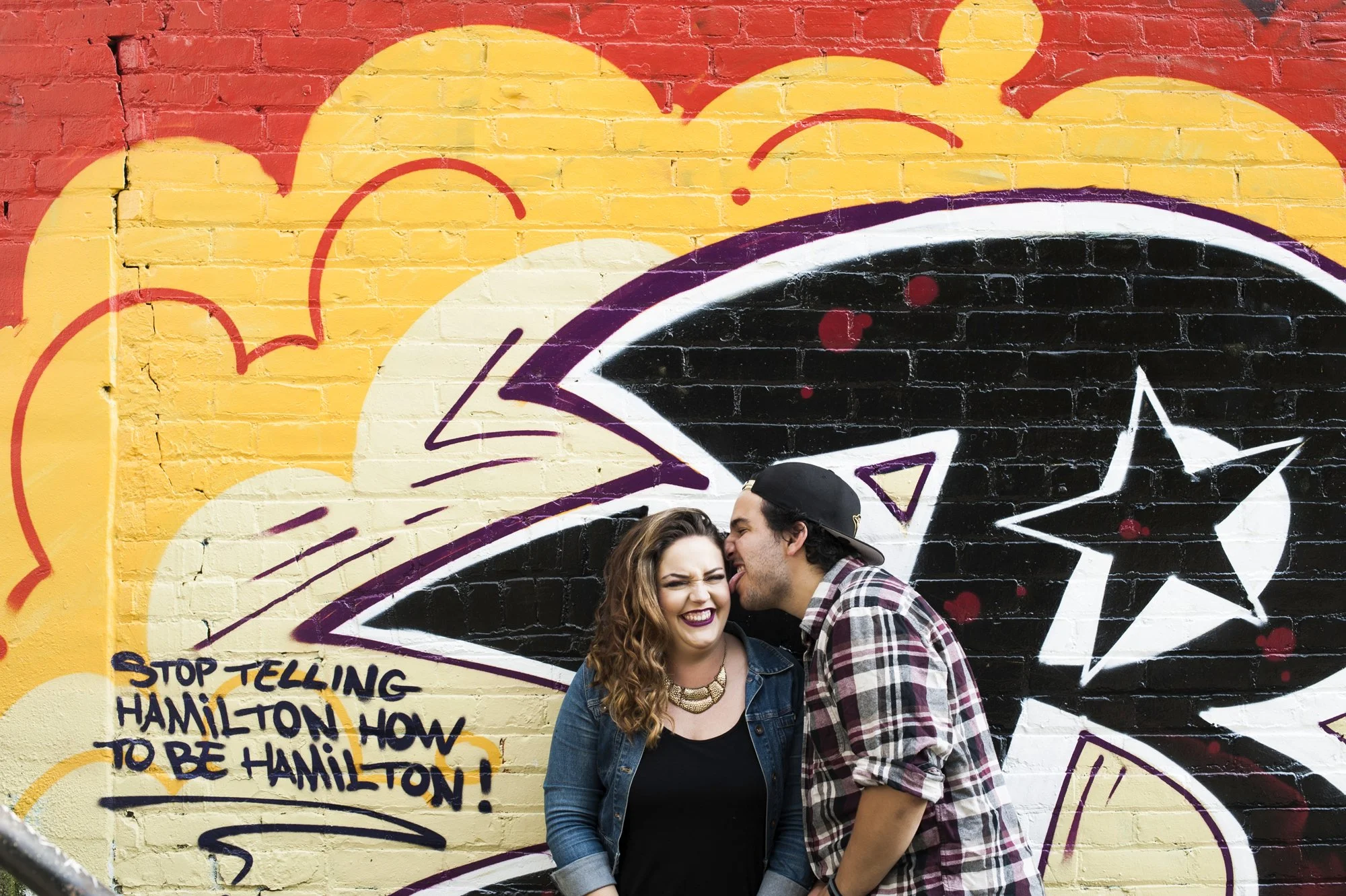 A young couple standing close to each other in front of a colorful graffiti wall, the man whispering in the woman's ear and both smiling.
