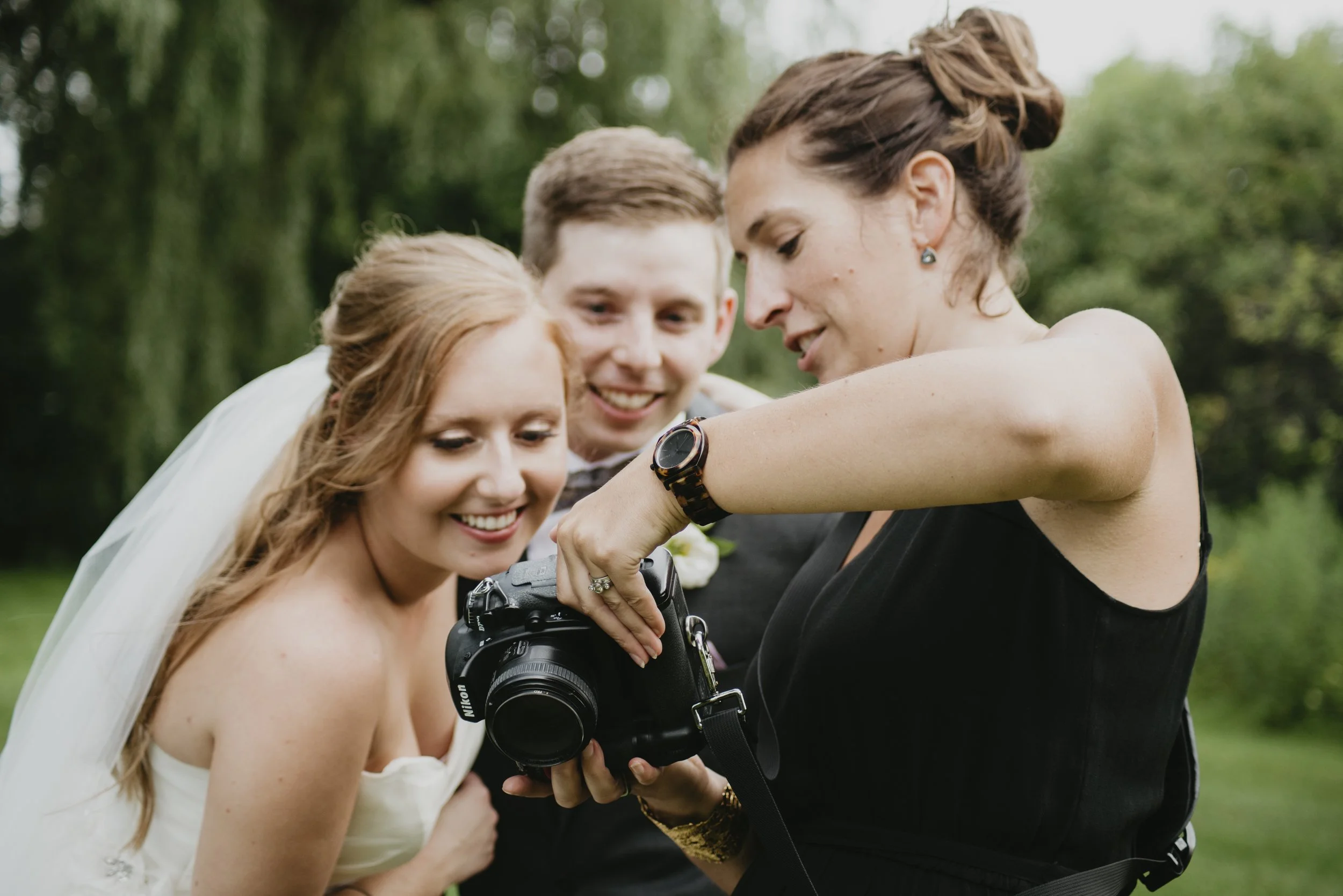 A bride with long red hair and a white gown is smiling as she looks at a camera held by a woman, while a man looks on. The woman holding the camera is wearing black and has short hair, and the man is dressed in a formal suit. They are outdoors with green trees in the background.
