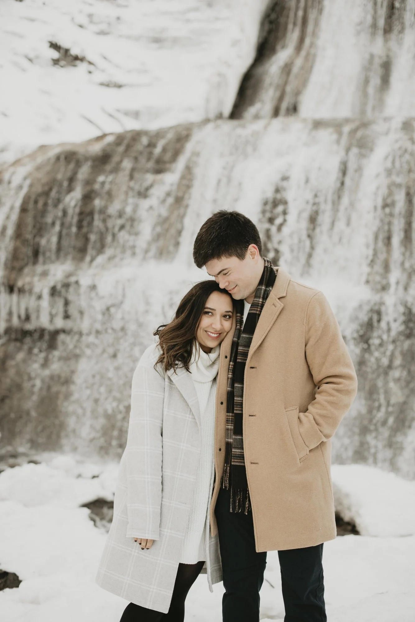 A young couple standing close together outdoors in a snowy landscape. The woman has long dark hair and is smiling, wearing a white coat and black tights. The man has short dark hair, is smiling, and is wearing a tan coat and a scarf. Behind them is a