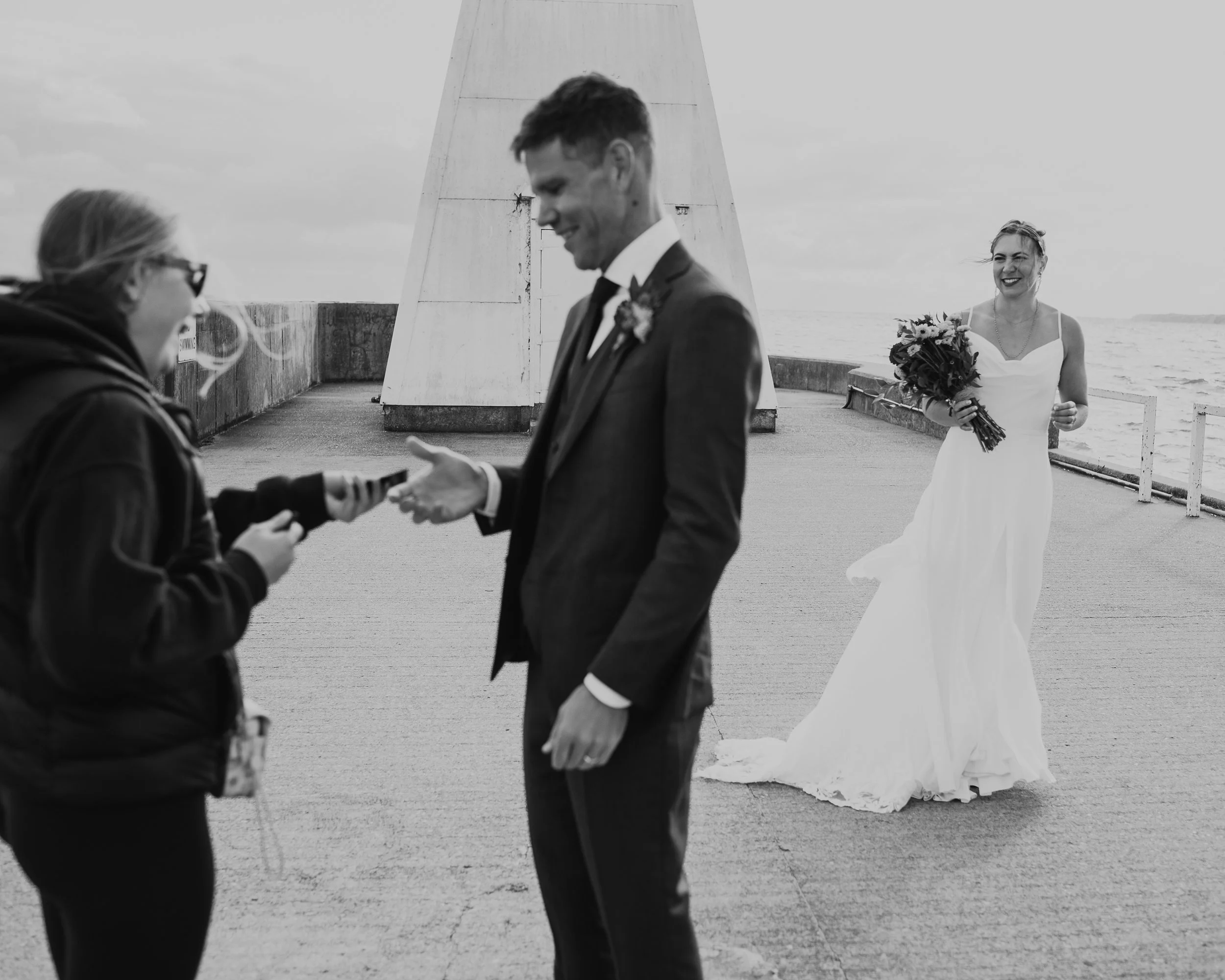 A bride in a white wedding dress holding a bouquet, smiling as she approaches a groom in a dark suit who is receiving a ring from a woman in casual clothing during an outdoor wedding ceremony near the ocean.