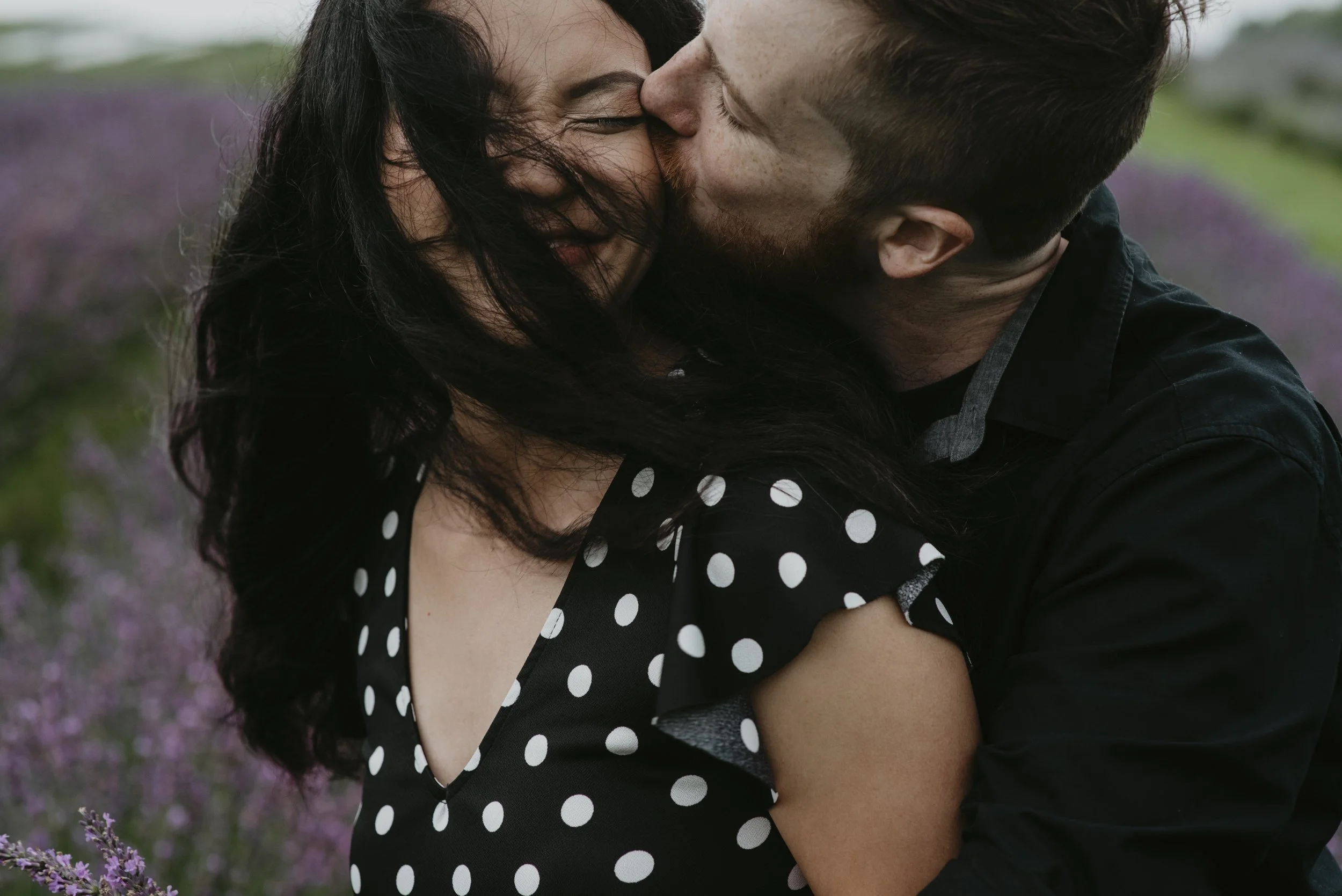 A couple embracing outdoors, the woman with dark hair and a polka dot dress smiling as they kiss, with blurred purple flowers in the background.
