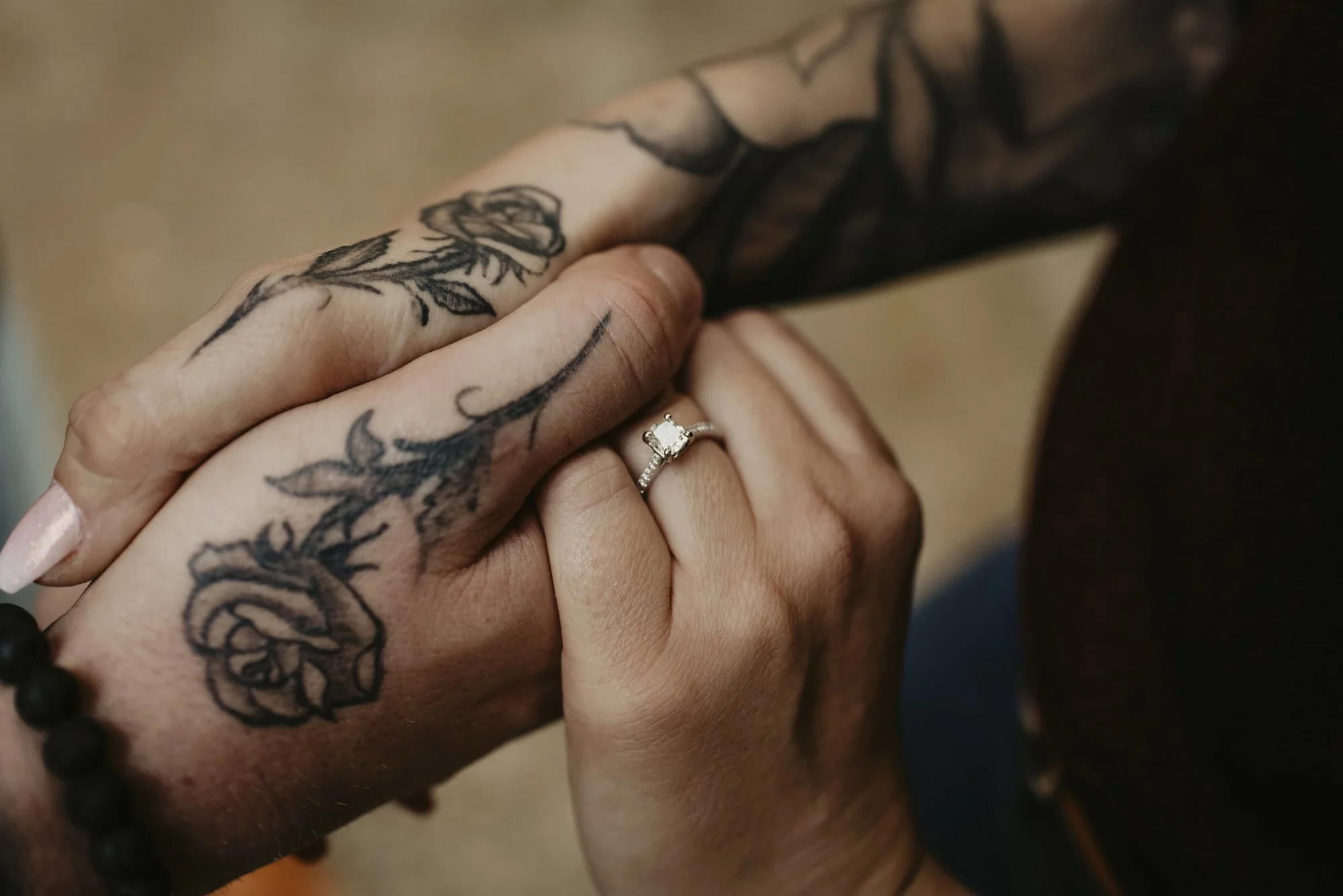 Close-up of two hands holding each other, one tattooed with a floral design and wearing a silver engagement ring with a central diamond. The background is blurred.