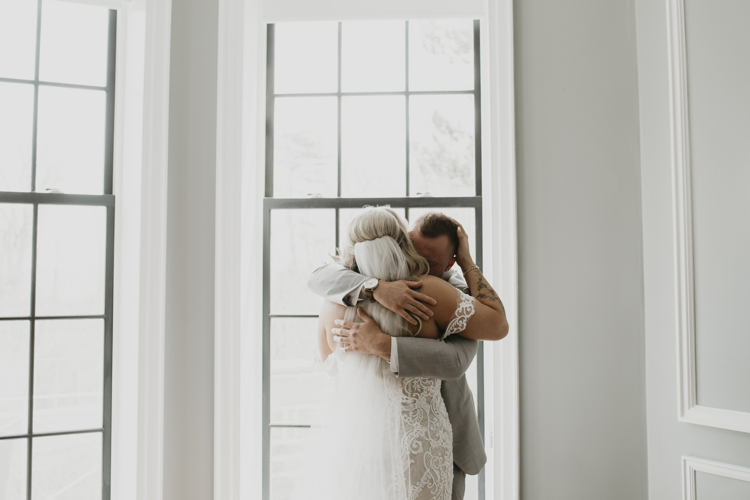A bride and groom hugging inside near large windows during a wedding celebration.