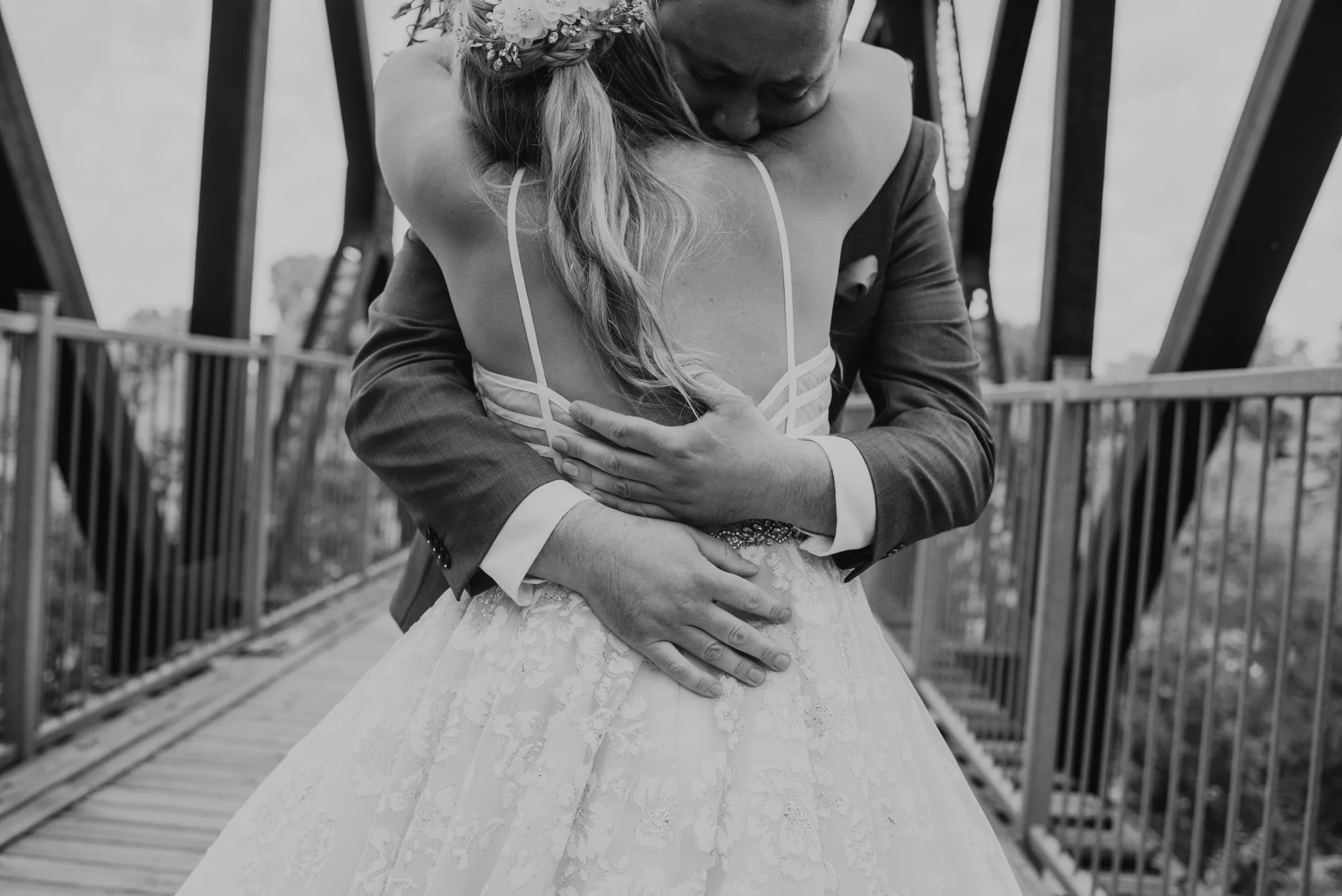 Black and white photo of a bride and groom hugging each other tightly on a bridge. The bride wears a wedding dress and a floral headpiece, and the groom wears a suit. The focus is on their embrace, with their faces obscured.