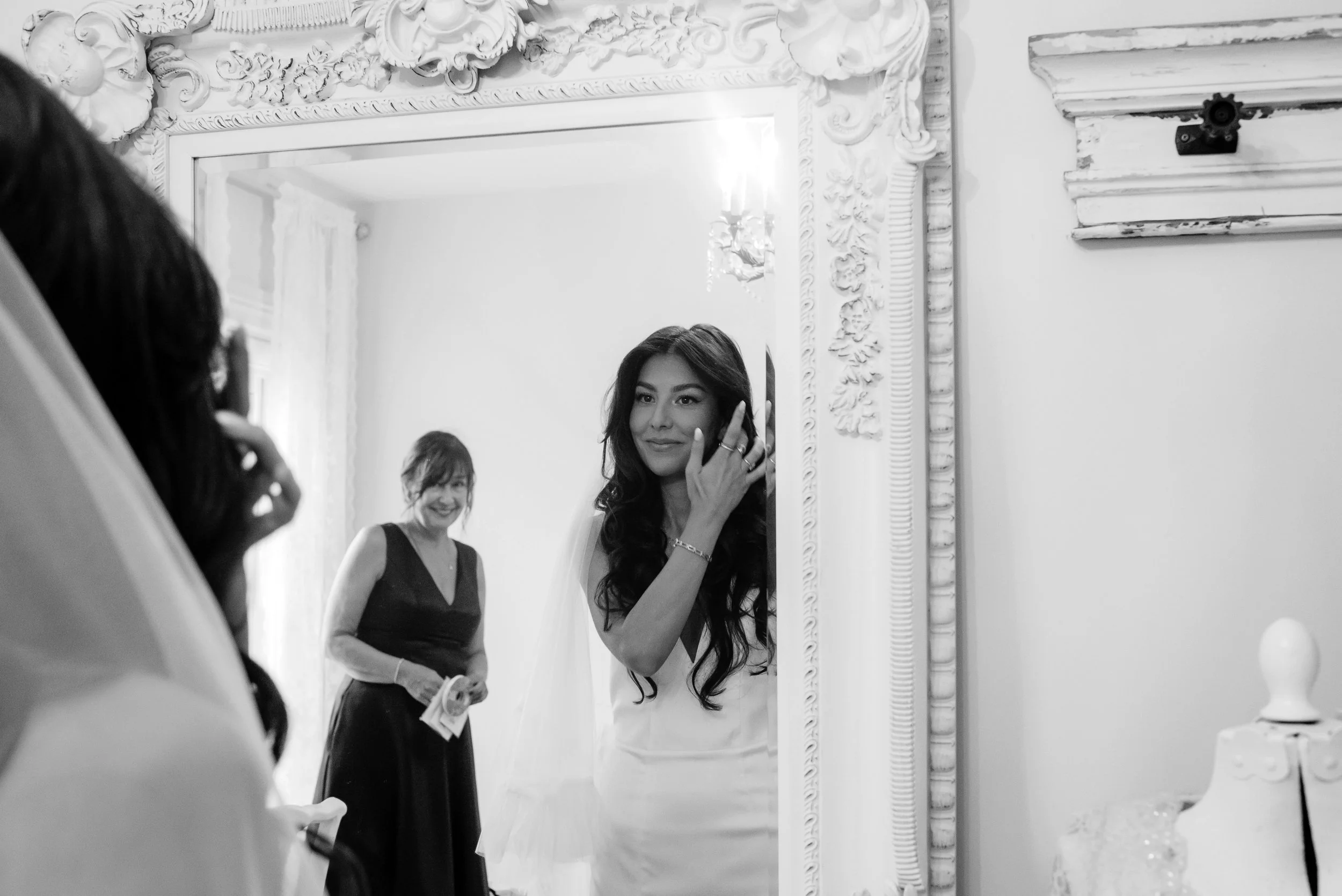 A woman with long dark hair in a white dress smiles at herself in a mirror, her hand touching her face, while another woman in a dark dress looks on and smiles in the background.