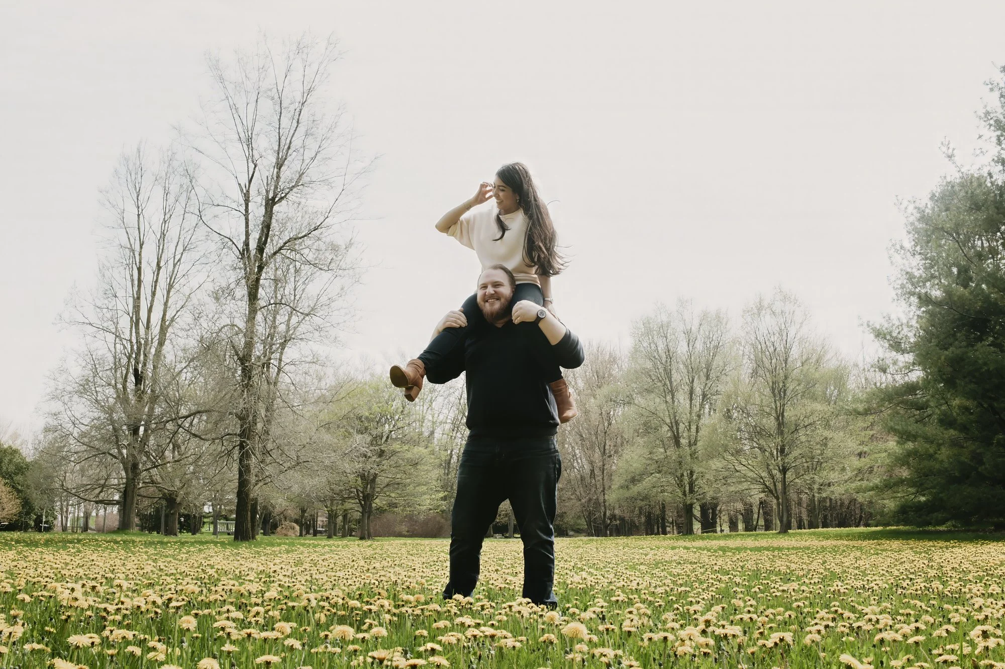 A man carrying a woman on his shoulders in a park with blooming flowers and trees, overcast sky.
