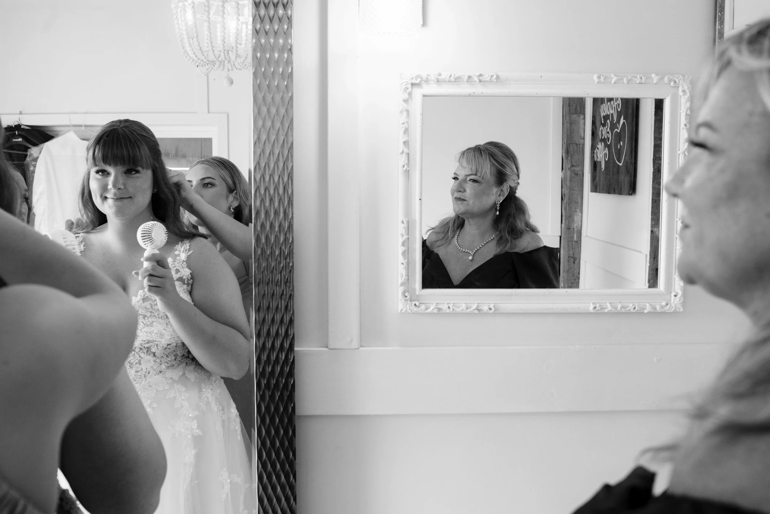 A young woman in a wedding dress getting her hair styled in a mirror, with two women watching, one older woman with jewelry and a black dress, and another woman partially visible on the right.