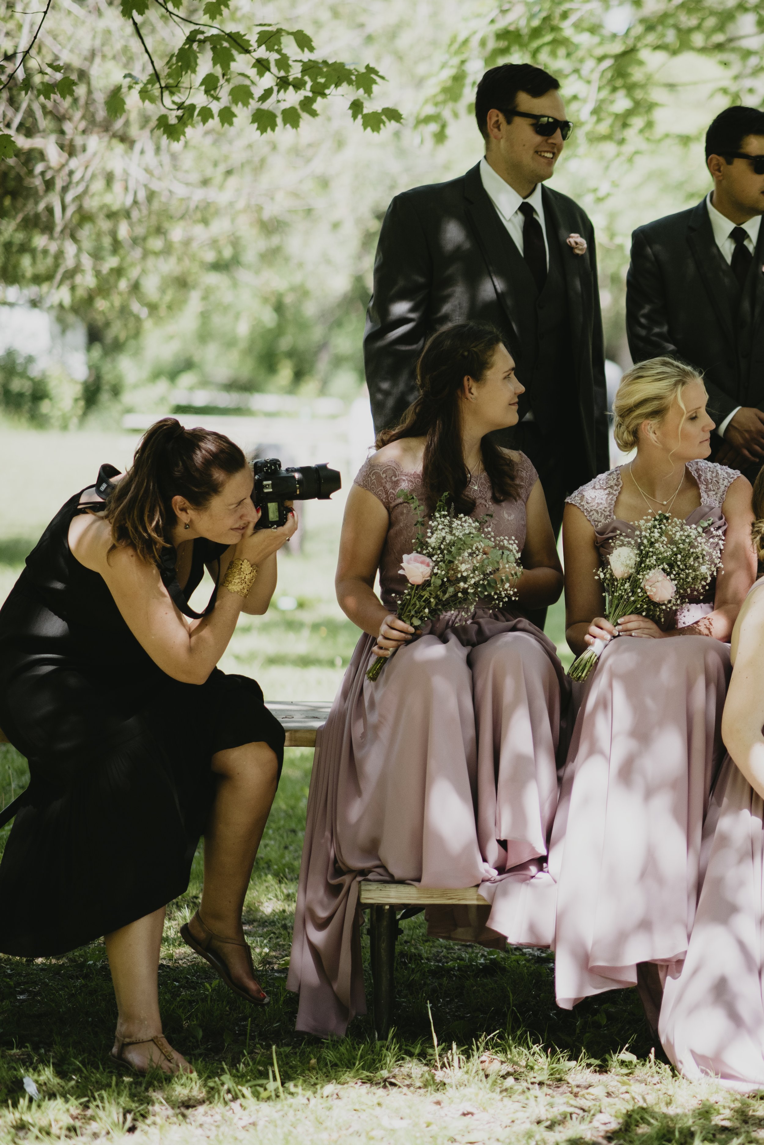 Photographer crouched taking picture of women at outdoor wedding ceremony with groomsmen standing behind.