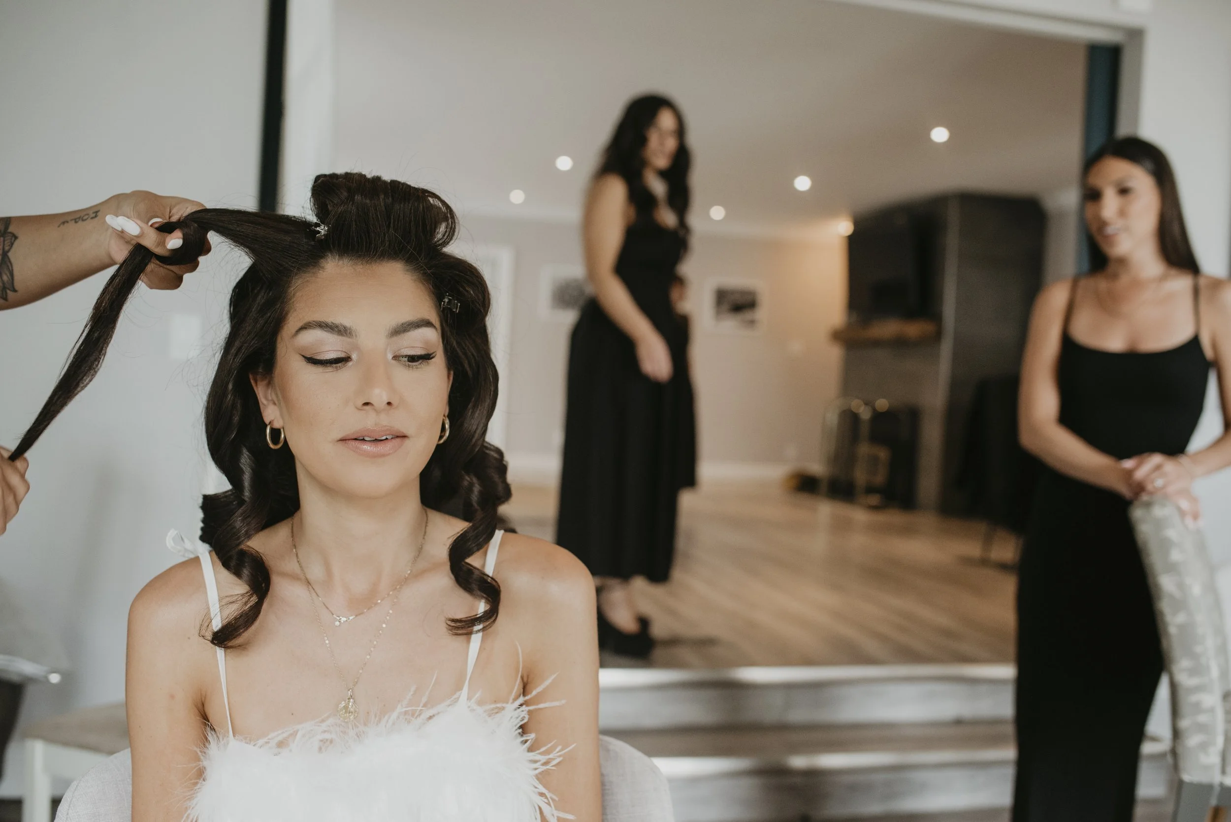 A woman getting her hair styled in a salon, with two other women in black dresses in the background.