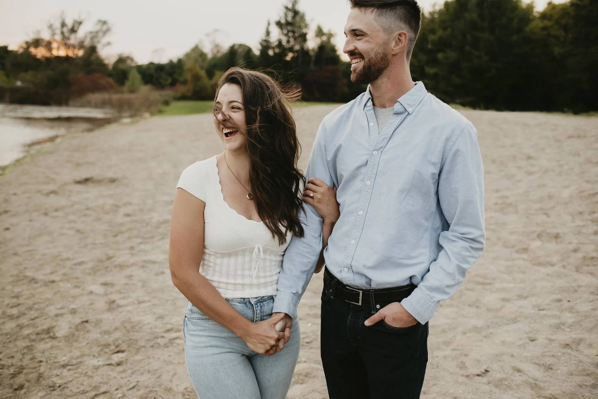 A happy couple holding hands and smiling on a sandy beach during sunset, with trees and a cloudy sky in the background.