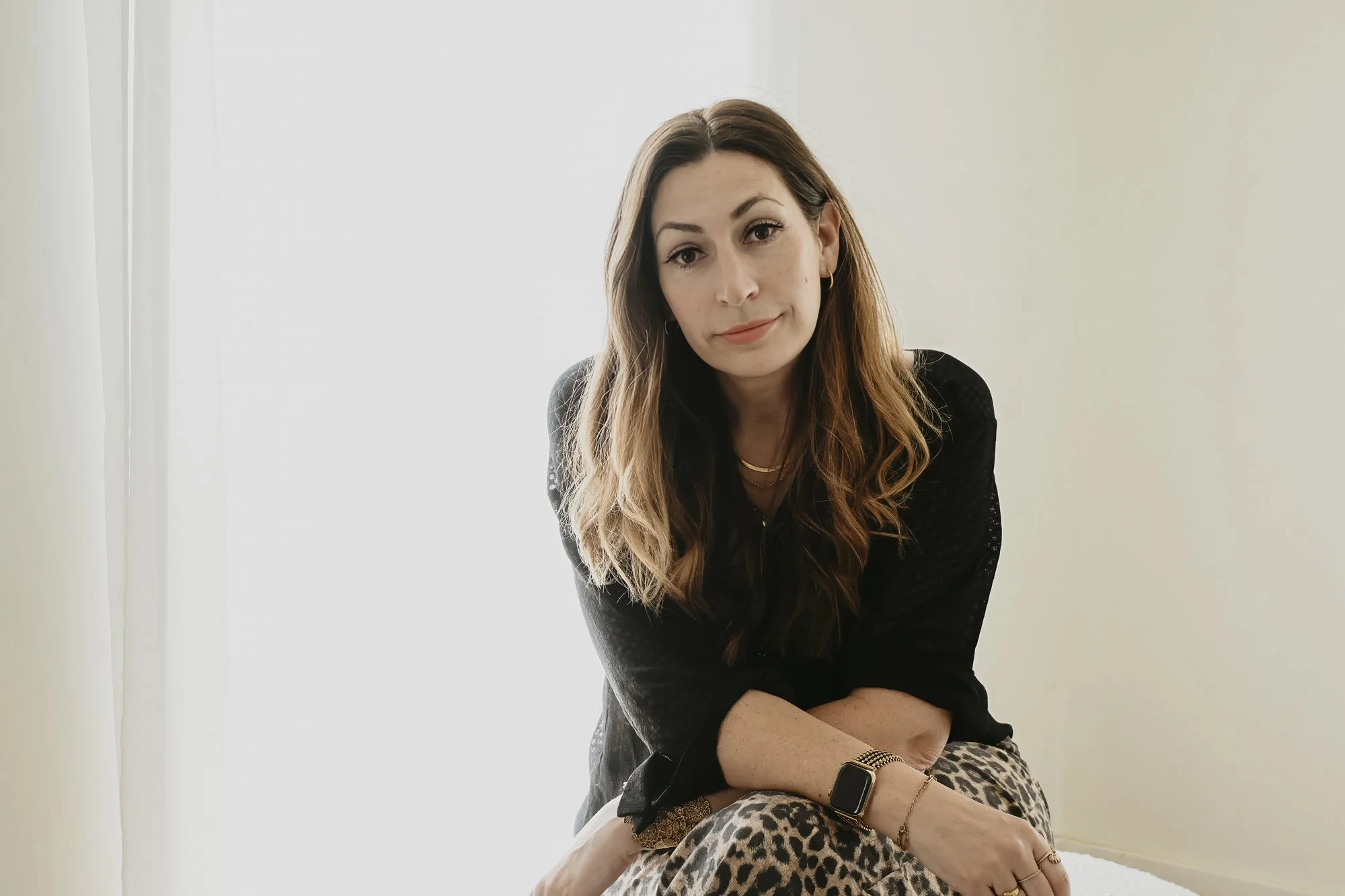 A woman with long brown hair and light makeup sitting in a well-lit room with plain white and beige walls, wearing a black top, leopard print pants, a smartwatch, and gold jewelry.
