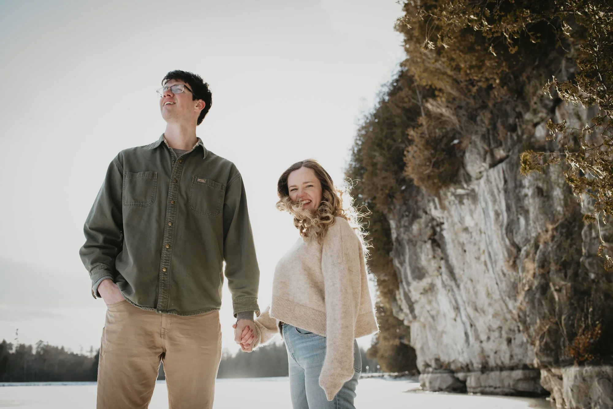 A young couple holding hands and smiling outdoors, standing near a rocky cliff with autumn foliage on a cloudy day.