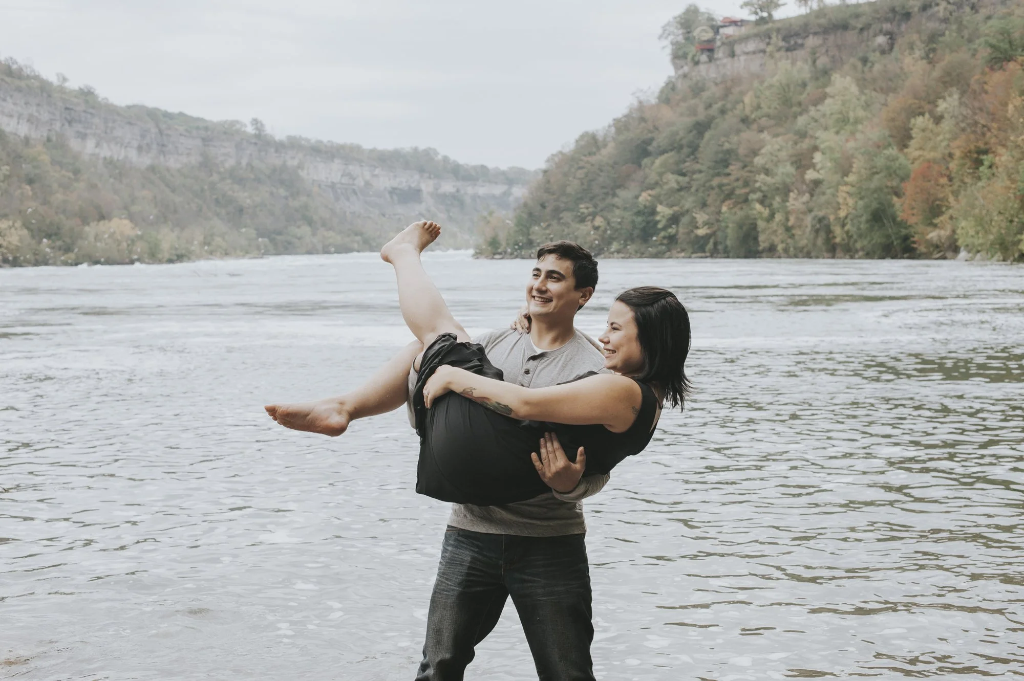 A man is holding a woman in his arms while standing in a river. They are smiling and looking at each other. The background features a scenic view of hills with autumn-colored trees and a cloudy sky.