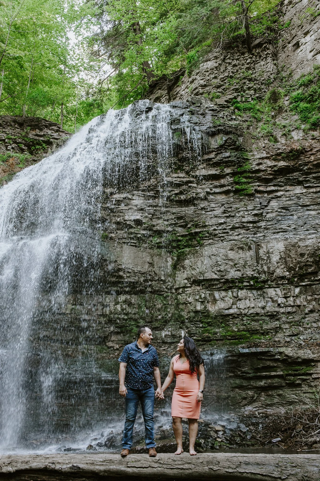 A couple holding hands and standing on a rock in front of a waterfall surrounded by green trees.