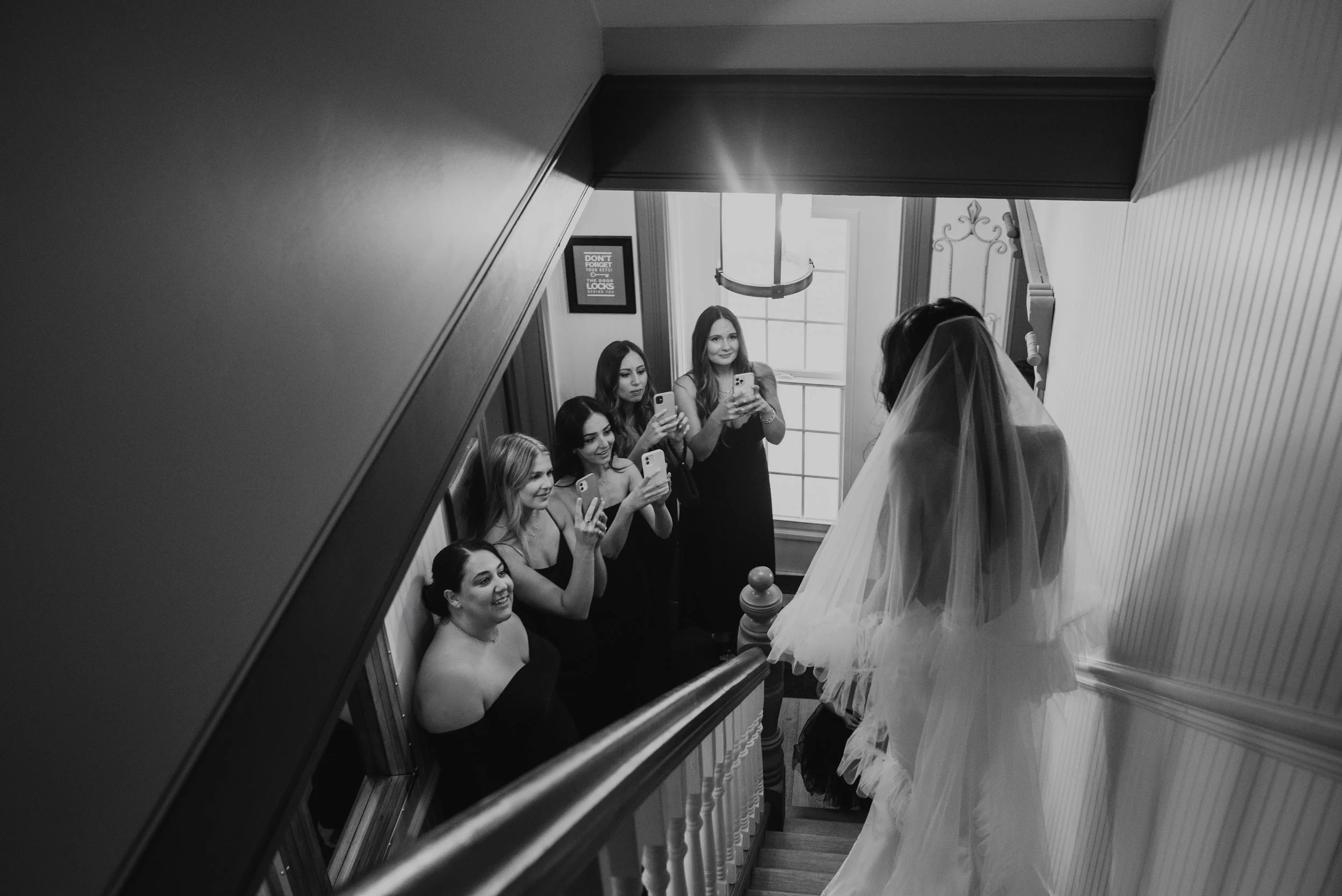 A woman in a wedding dress with a veil standing on a staircase, surrounded by five women taking photos of her with their phones. The setting appears to be indoors, with natural light coming through a large window in the background.