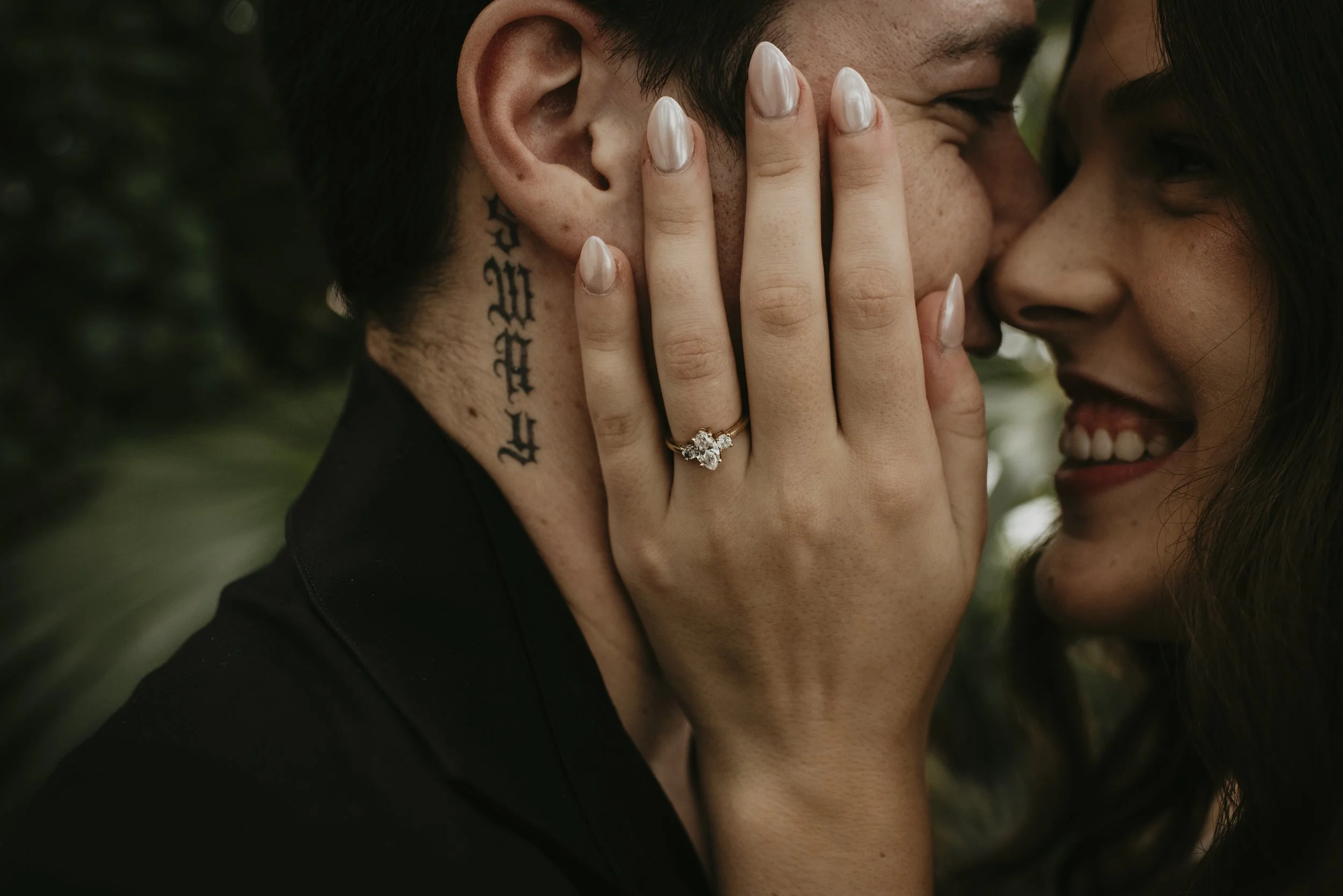 A couple close together, with the man whispering into the woman's ear, her hand with a ring covering part of his face, showcasing affection and intimacy.