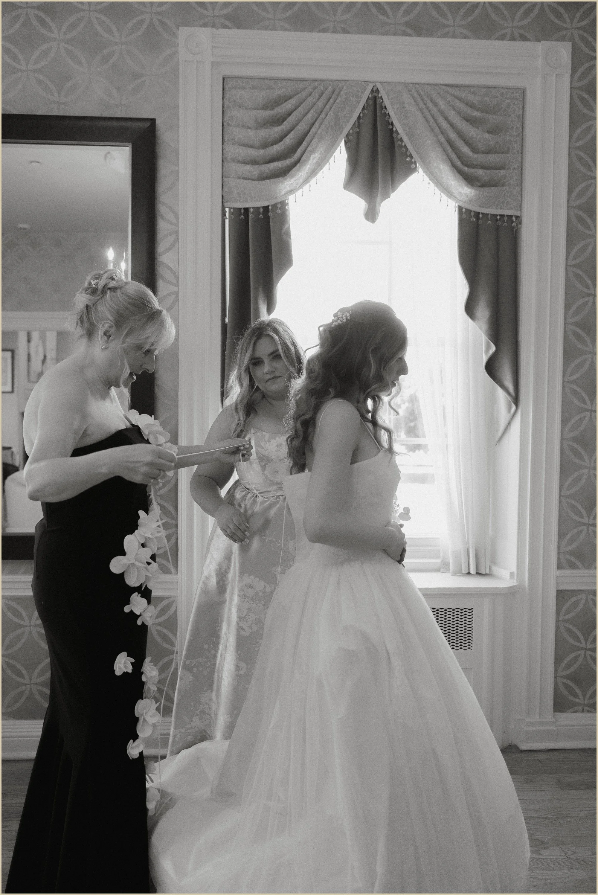 candid black and white photo of mother and sister helping bride with her dress