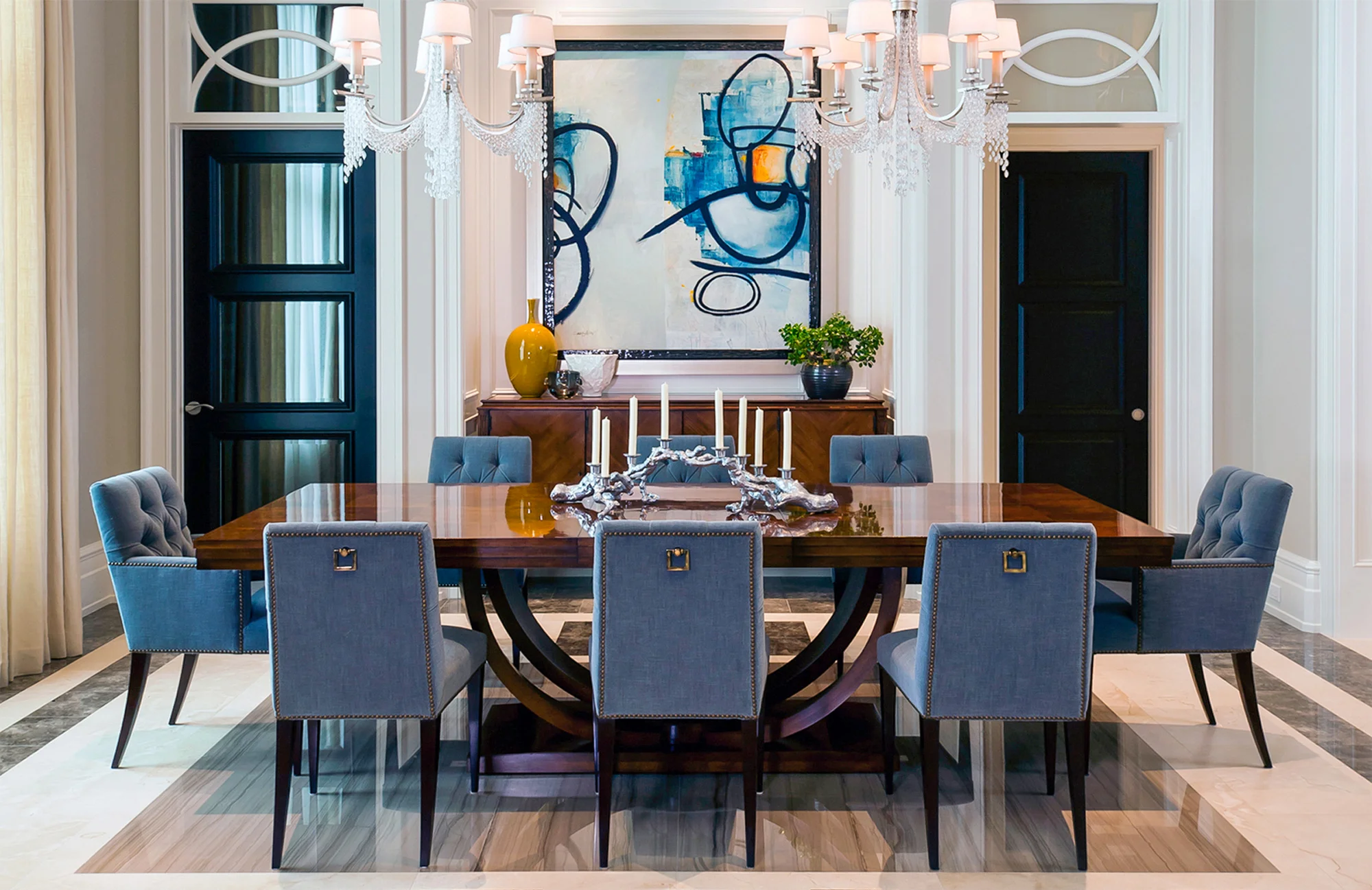 The natural tone of the dining table is complimented by bursts of sparkle, blue velvet chairs and artwork and a natural toned floor.