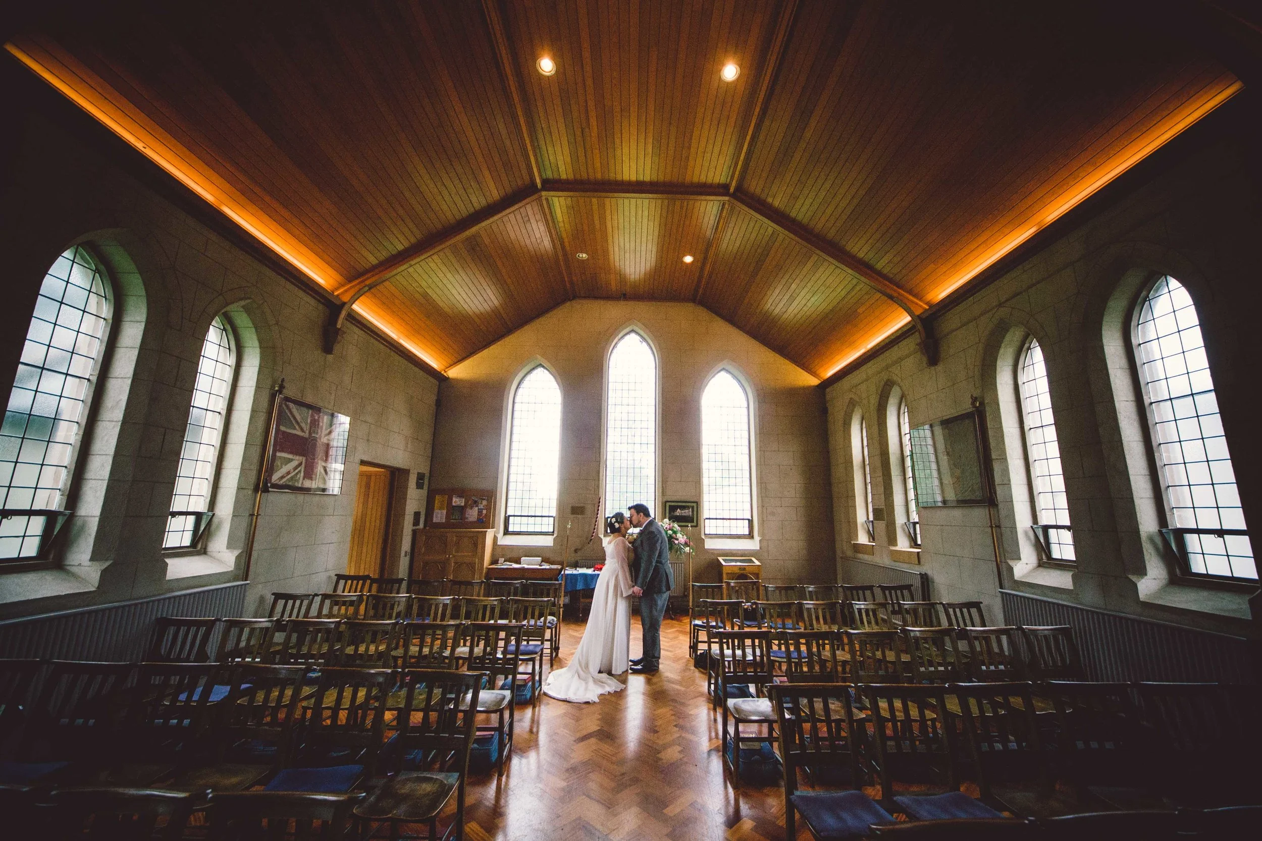 A bride and groom share a kiss inside a small chapel with tall, arched windows and a wooden ceiling.