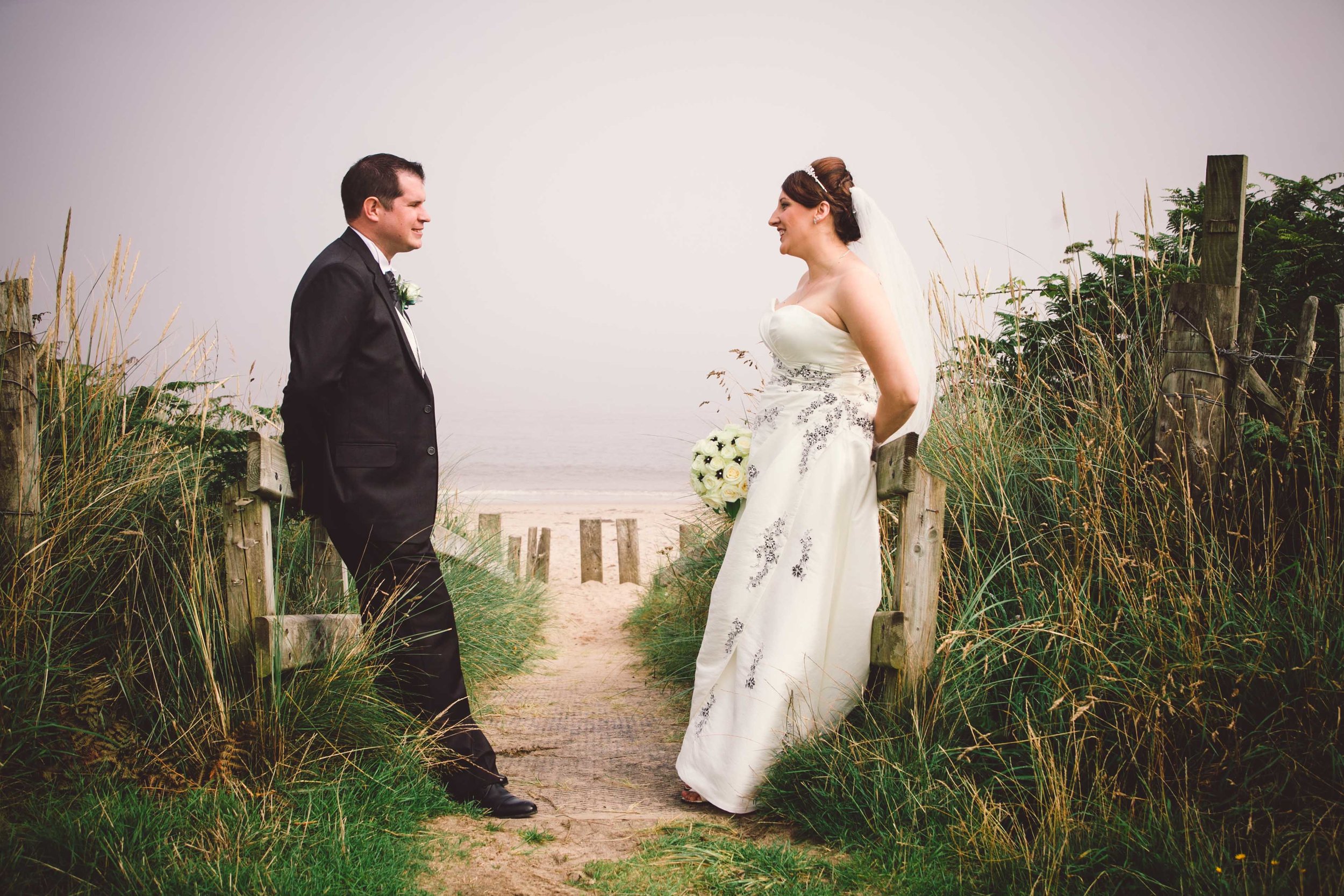 A bride and groom standing on a sandy path, facing each other, outdoors near the beach with beach grass and wooden posts surrounding them.