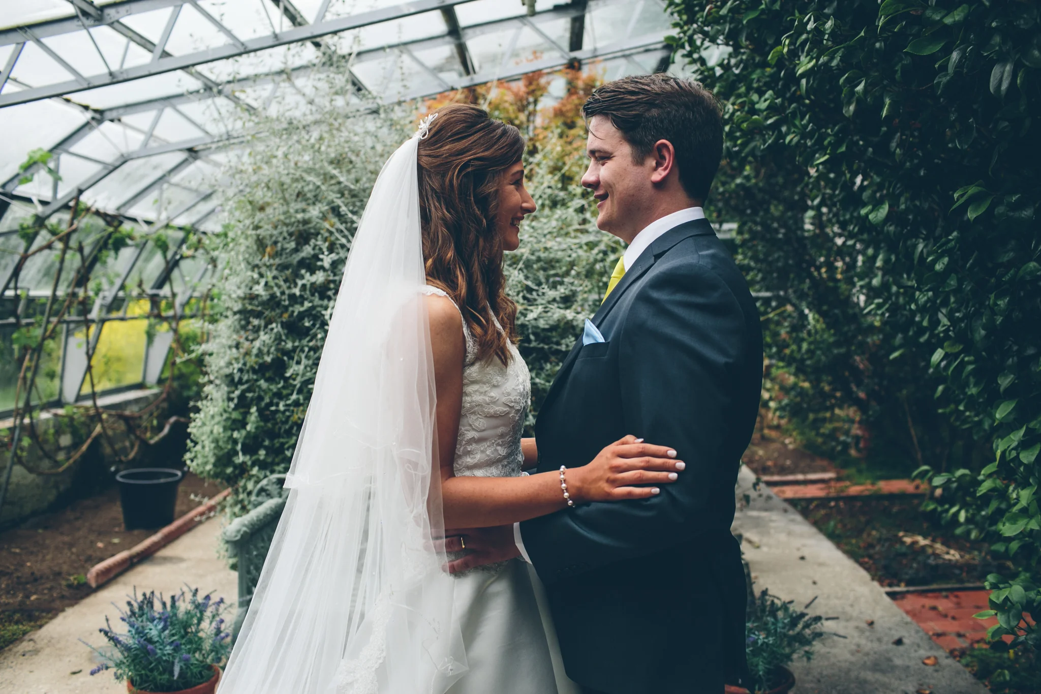 A bride and groom facing each other inside a greenhouse, smiling and holding each other gently. Larchfield Estate