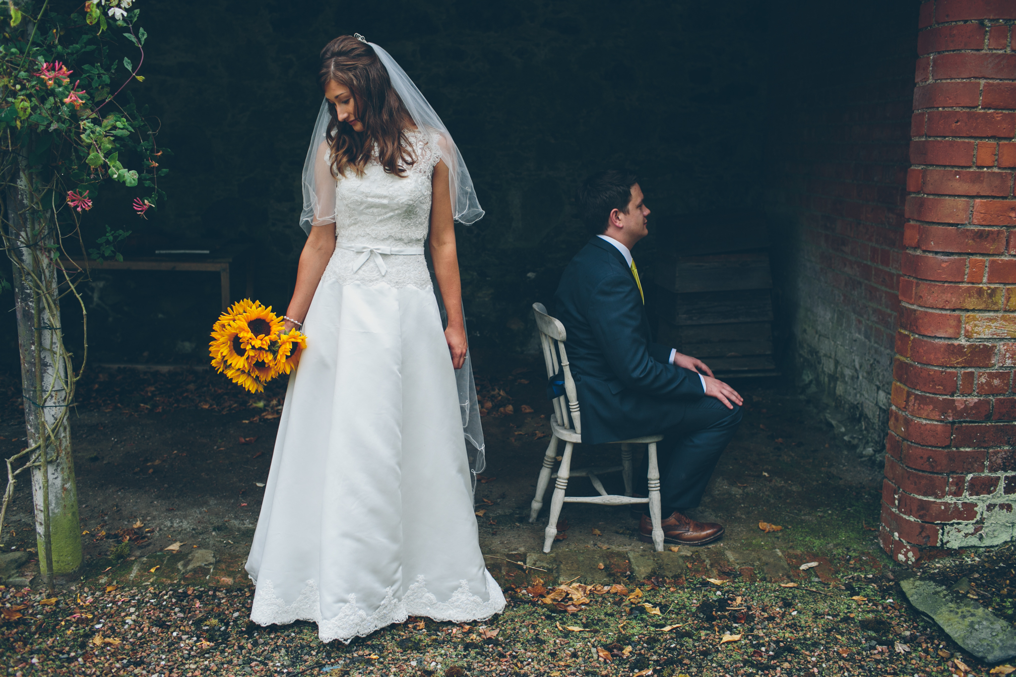 A bride in a white wedding dress with a veil holds a sunflower bouquet and looks down, standing outdoors near a brick wall. A man in a navy suit with a yellow tie sits on a white chair with his eyes closed, facing sideways, in a shaded area with gree
