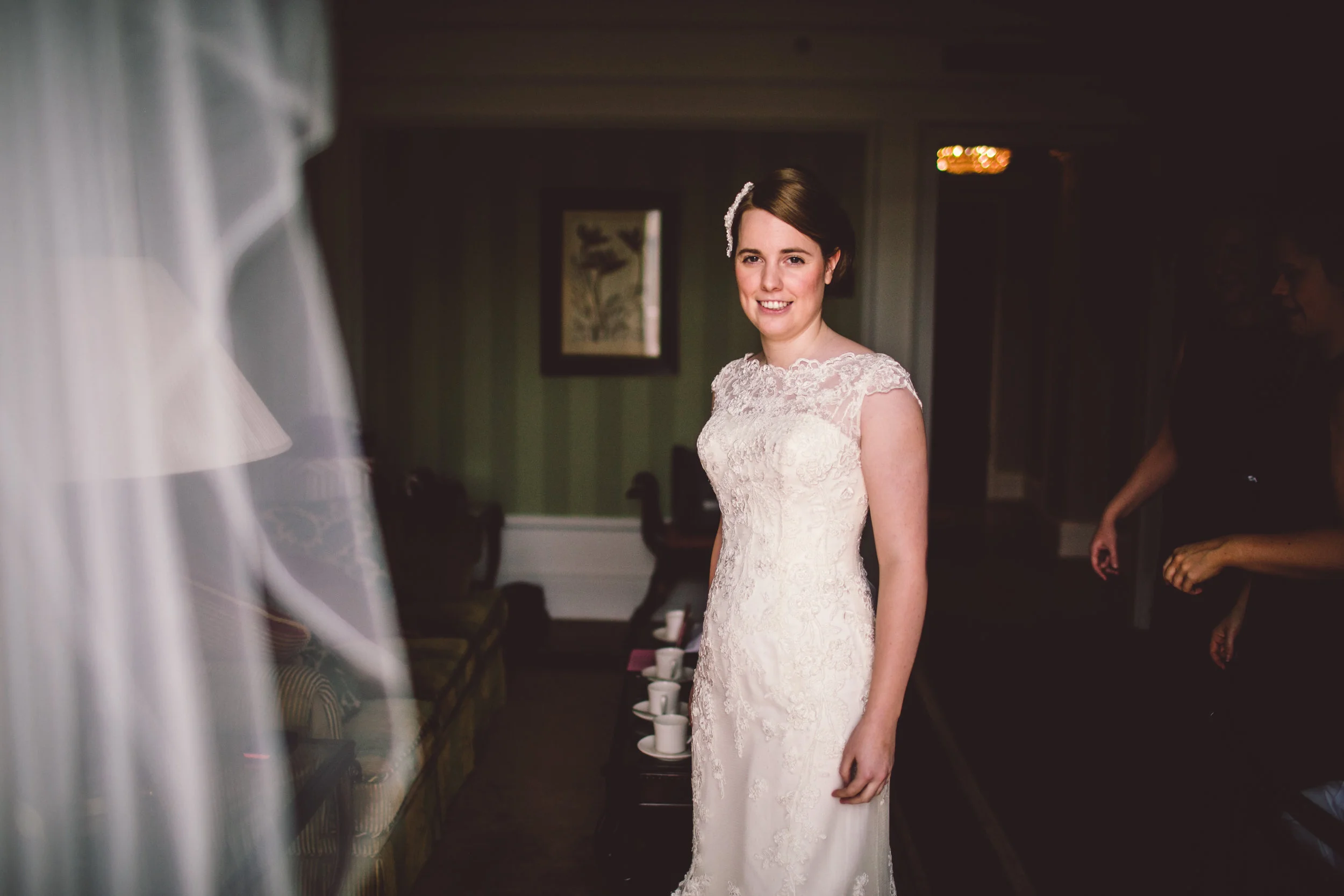 A smiling bride in a wedding dress with lace details, standing indoors in front of a green wall with framed artwork. A veil partially visible on the left side and couple of people in the background.
