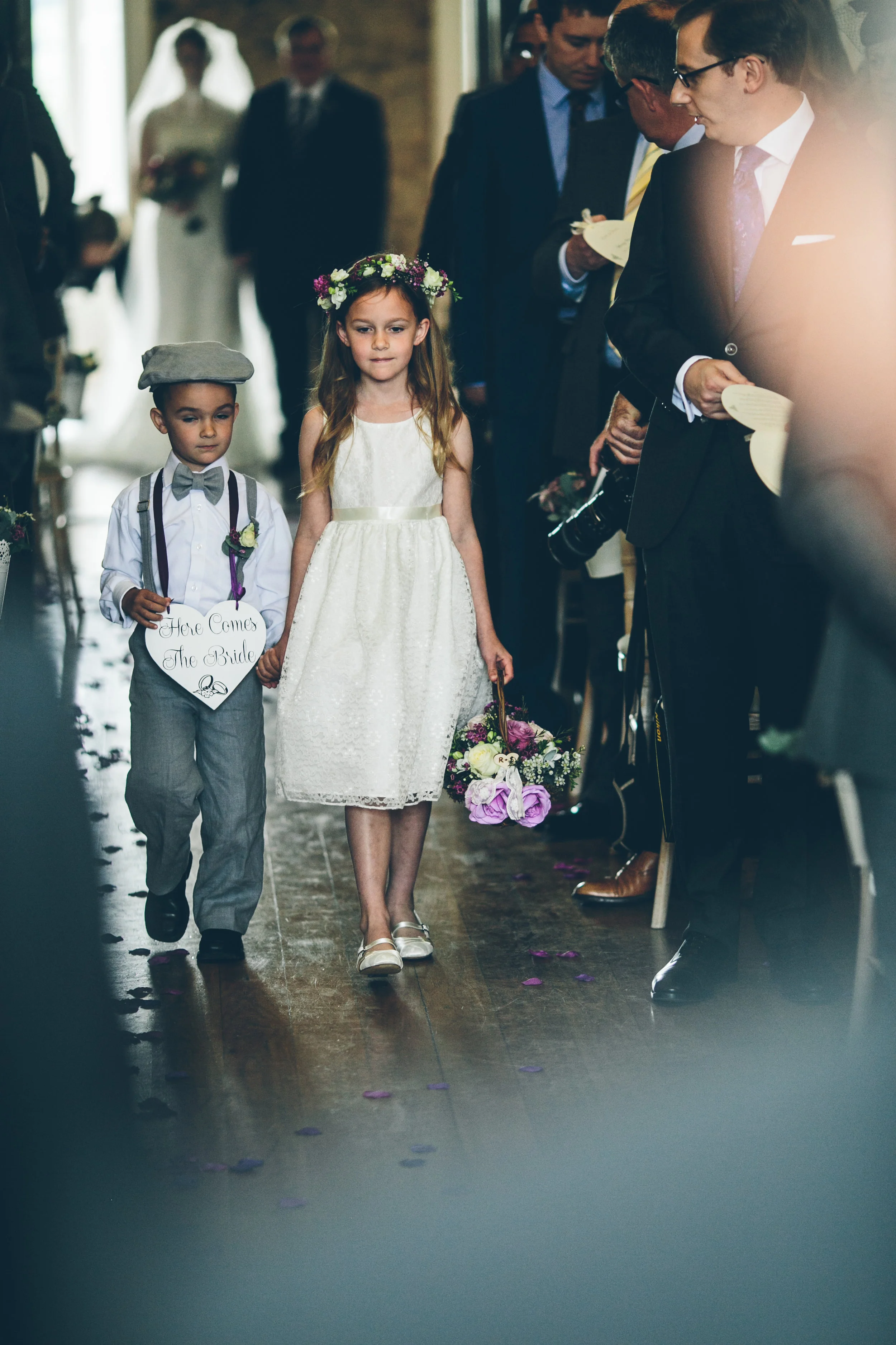 Children walking down aisle at a wedding ceremony, with a girl holding a basket and a boy holding a sign that says, 'Here Comes The Bride.'
