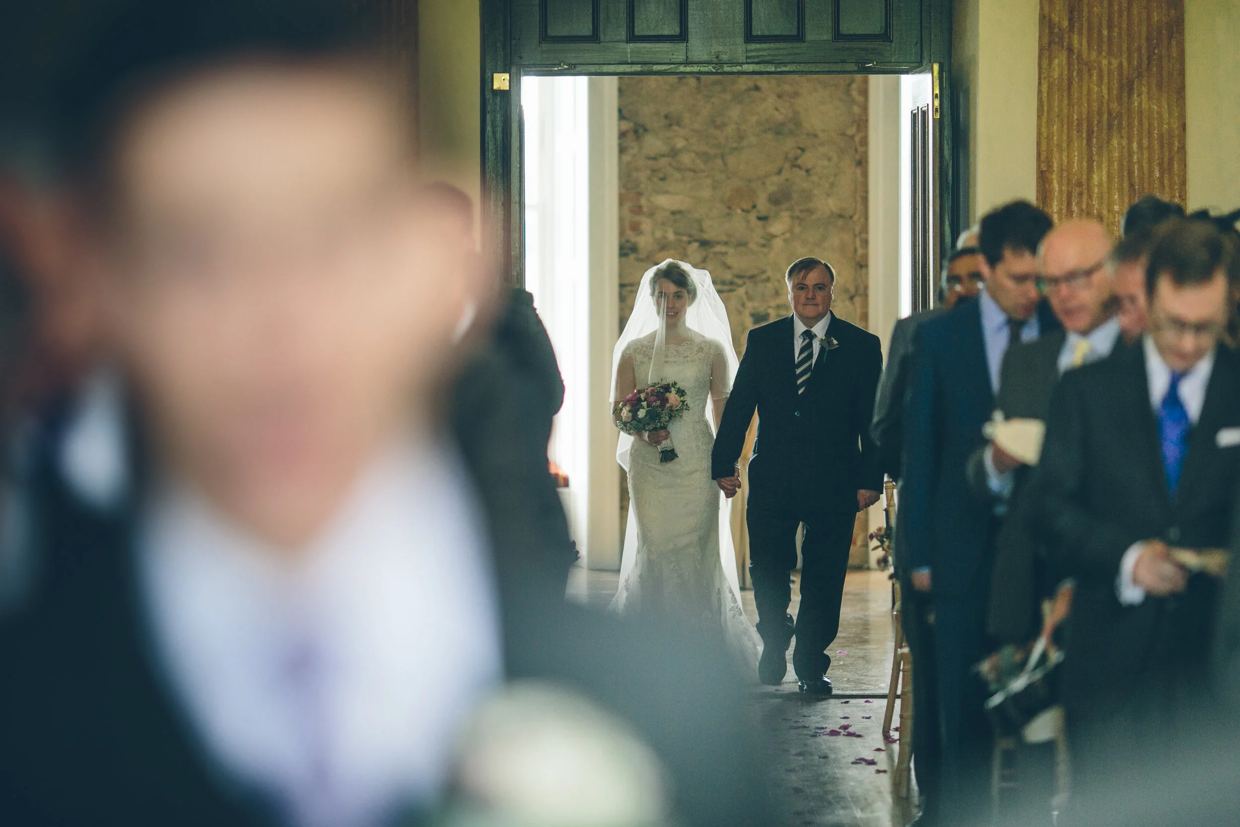 A bride in a white wedding dress and veil holding a bouquet walking down the aisle of a church, accompanied by a man in a suit, while guests are seated on both sides of the aisle.
