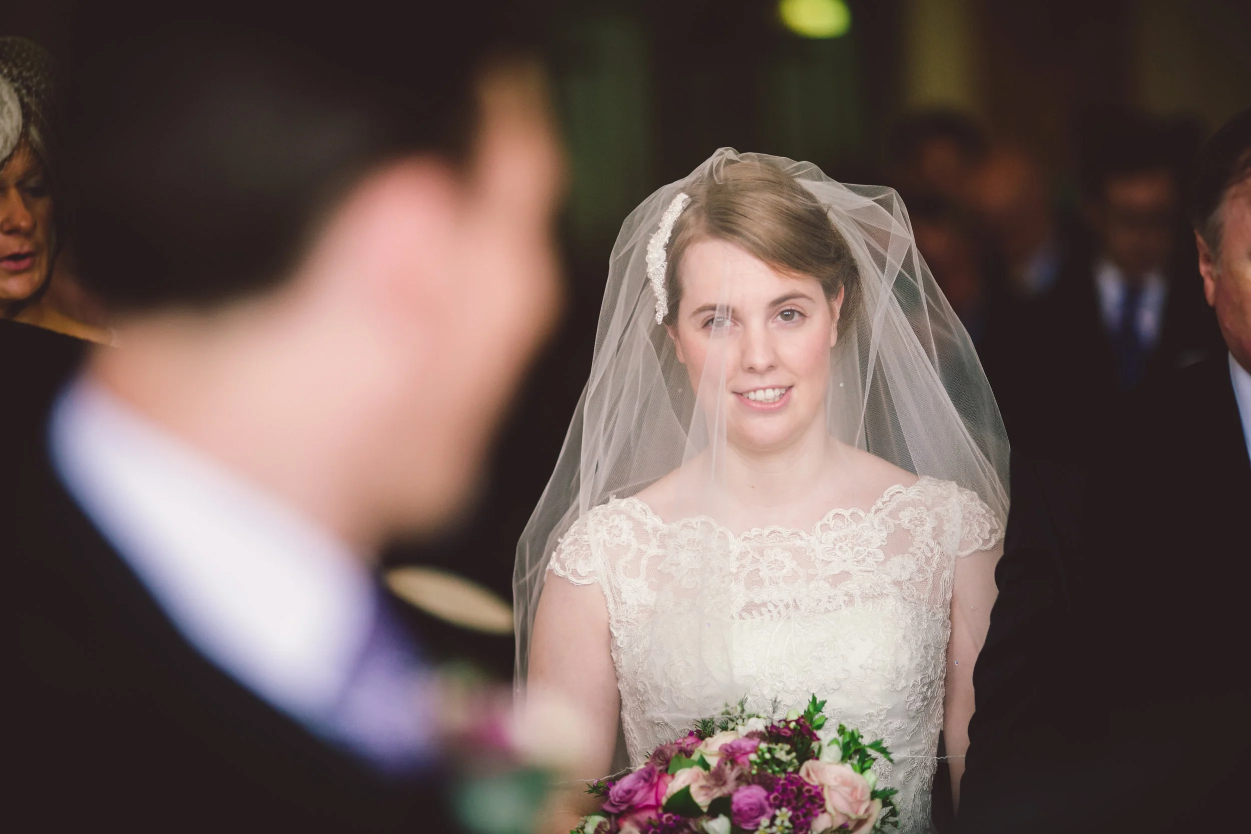 A bride wearing a lace wedding dress and veil, holding a bouquet of mixed flowers, smiling during her wedding ceremony, with guests in the background.