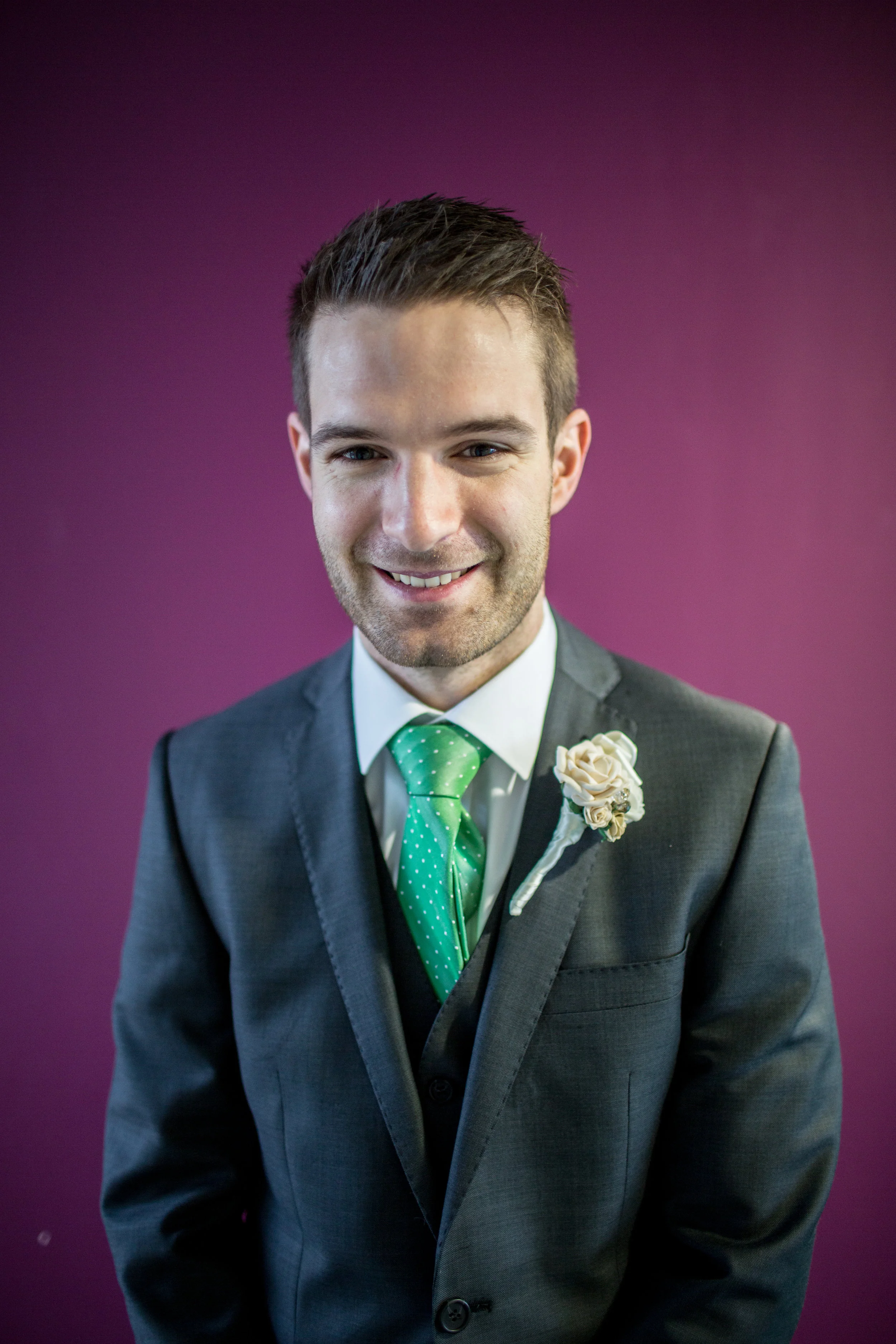 A smiling man in a gray suit, white shirt, and green tie with white dots, wearing a boutonnière, standing against a purple background.