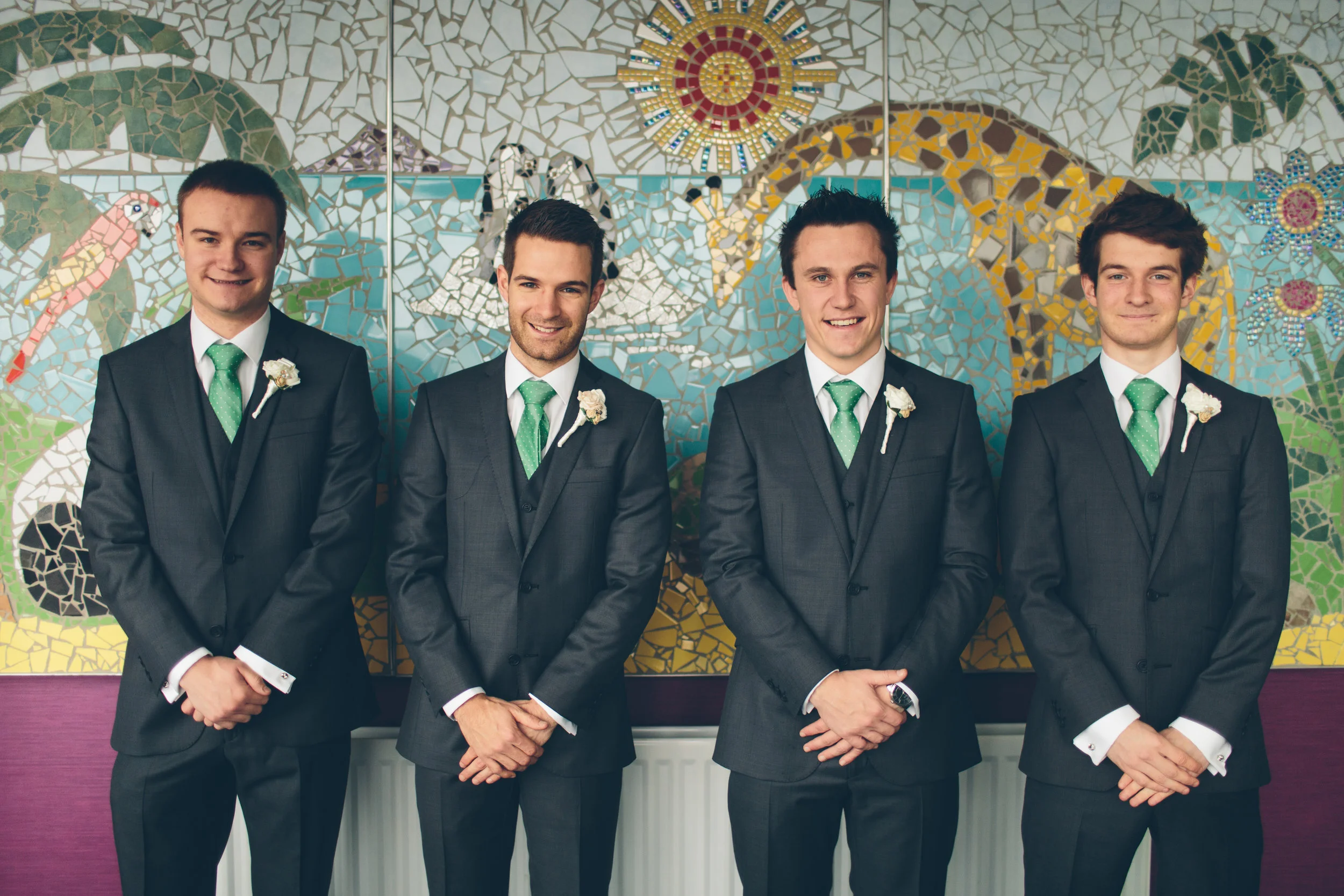 Four men dressed in dark suits with green ties, white shirts, and white boutonnieres on their left lapels, standing in front of a colorful mosaic wall with animal and nature designs.